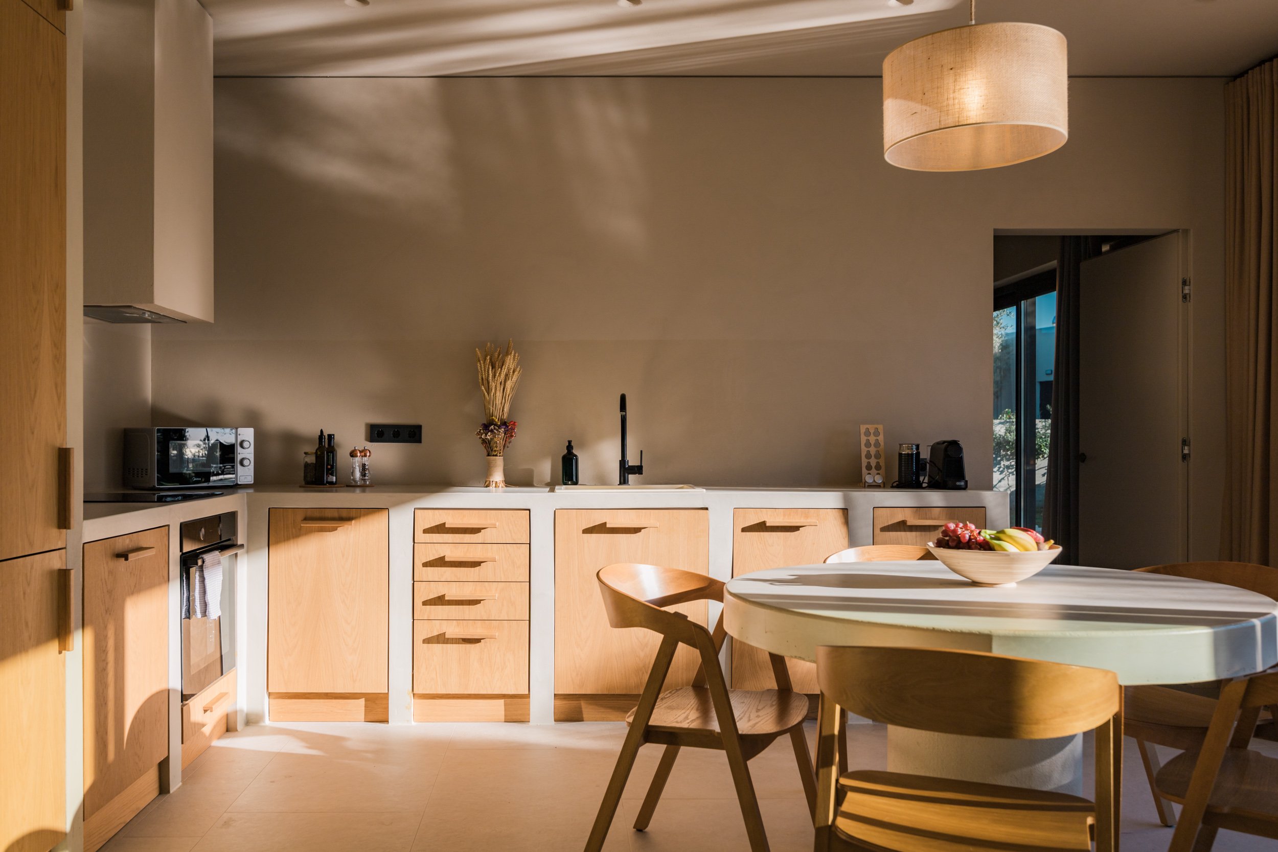 Kitchen with wooden cabinets, white countertop, black faucet, round dining table with wooden chairs, fruit bowl on the table, microwave, and large window with natural light.