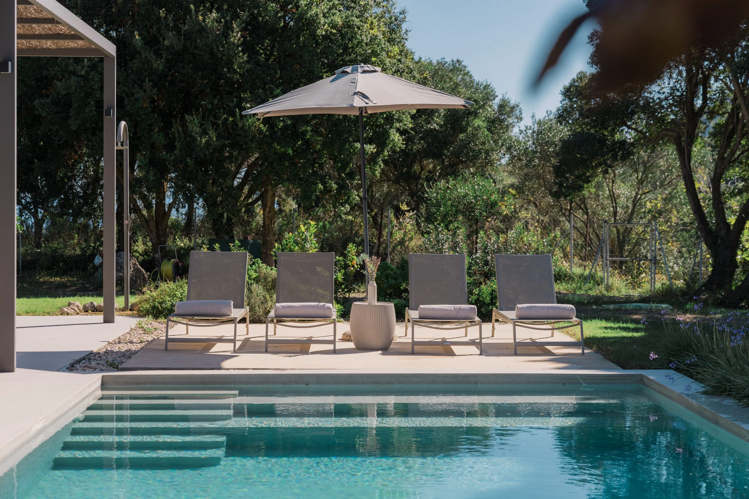 Poolside scene with four lounge chairs, a large umbrella, and a small table with a vase of flowers, set against a backdrop of trees and garden.