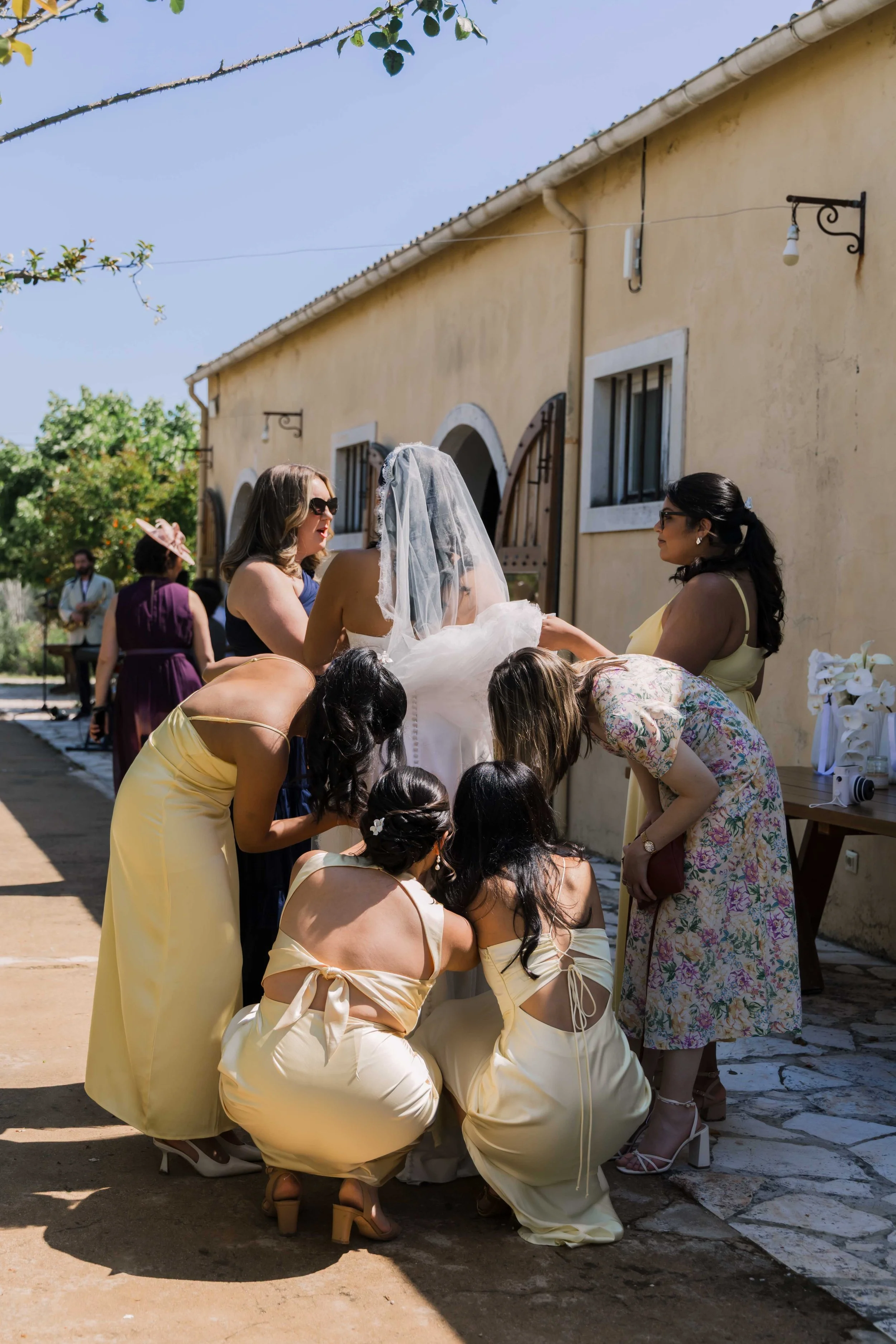 A bride surrounded by bridesmaids and guests at an outdoor wedding celebration. The bride wears a white wedding gown with a veil, and the bridesmaids and guests are dressed in yellow and floral dresses. They are engaged in a joyful moment outside a r
