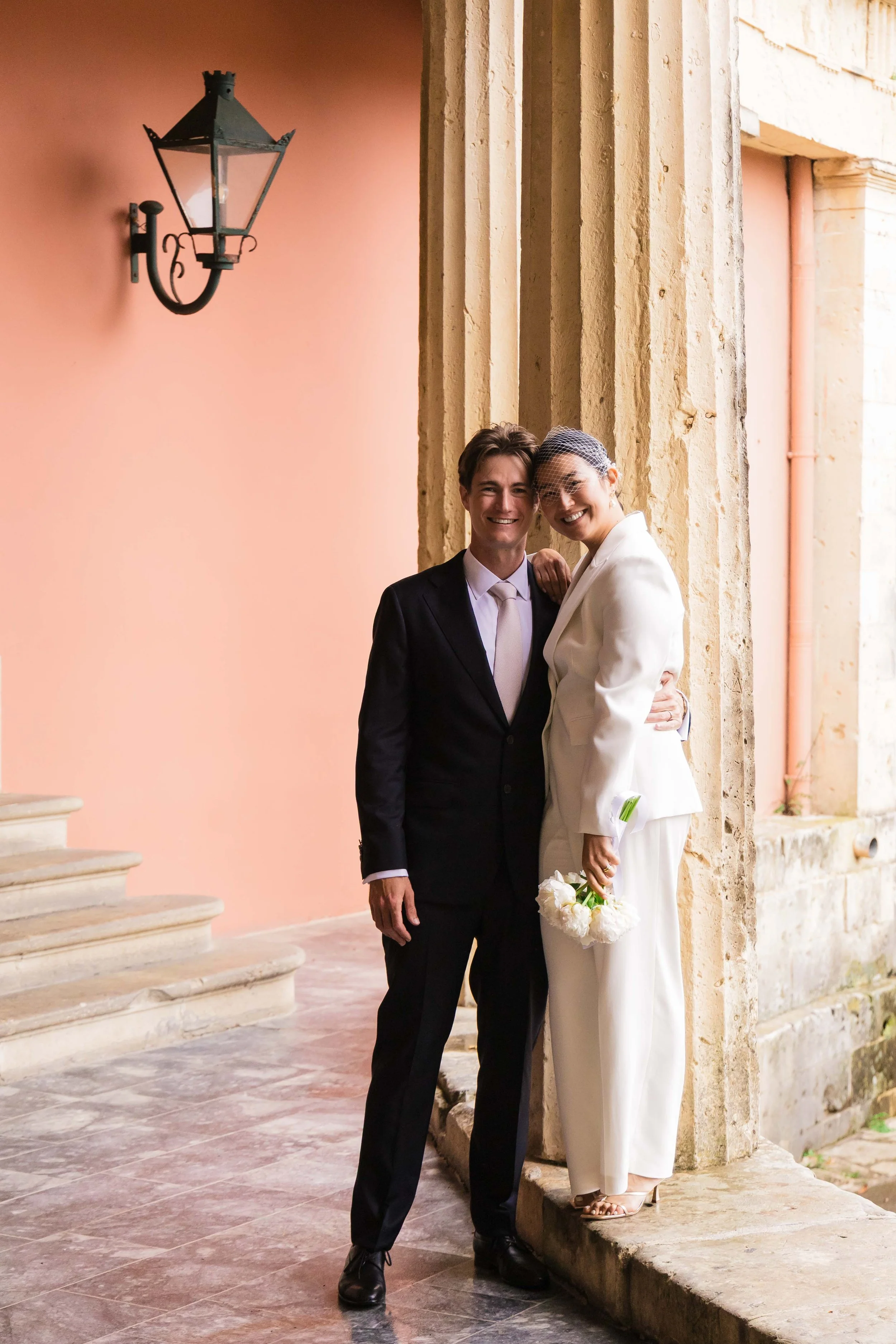 A bride and groom wearing wedding attire, smiling and standing close together against a stone archway in an outdoor setting.