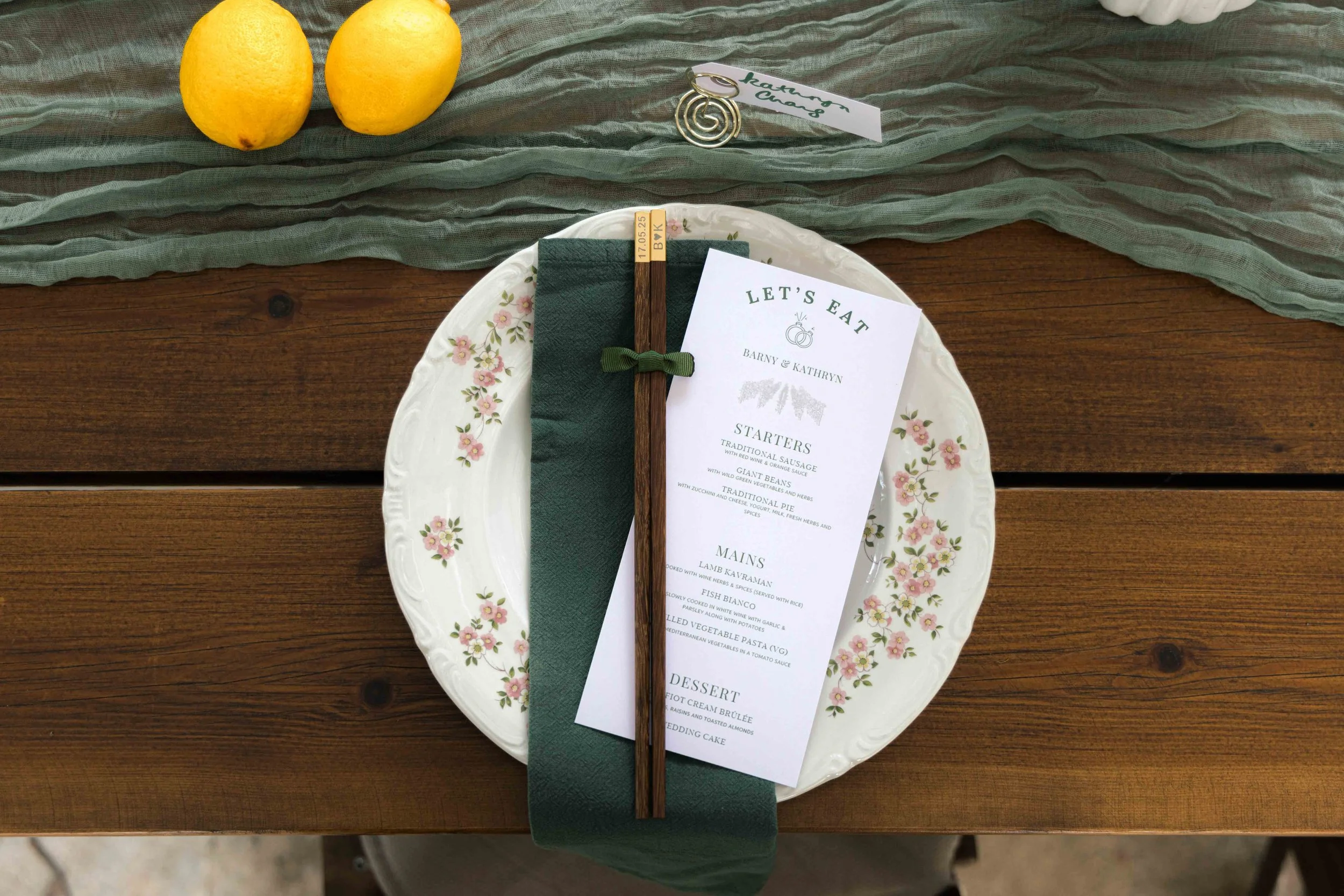 A table setting with a floral-patterned white plate, a green napkin, a small menu card, a pair of wooden chopsticks tied with a green ribbon, two lemons, and a small clip with a white and green label.