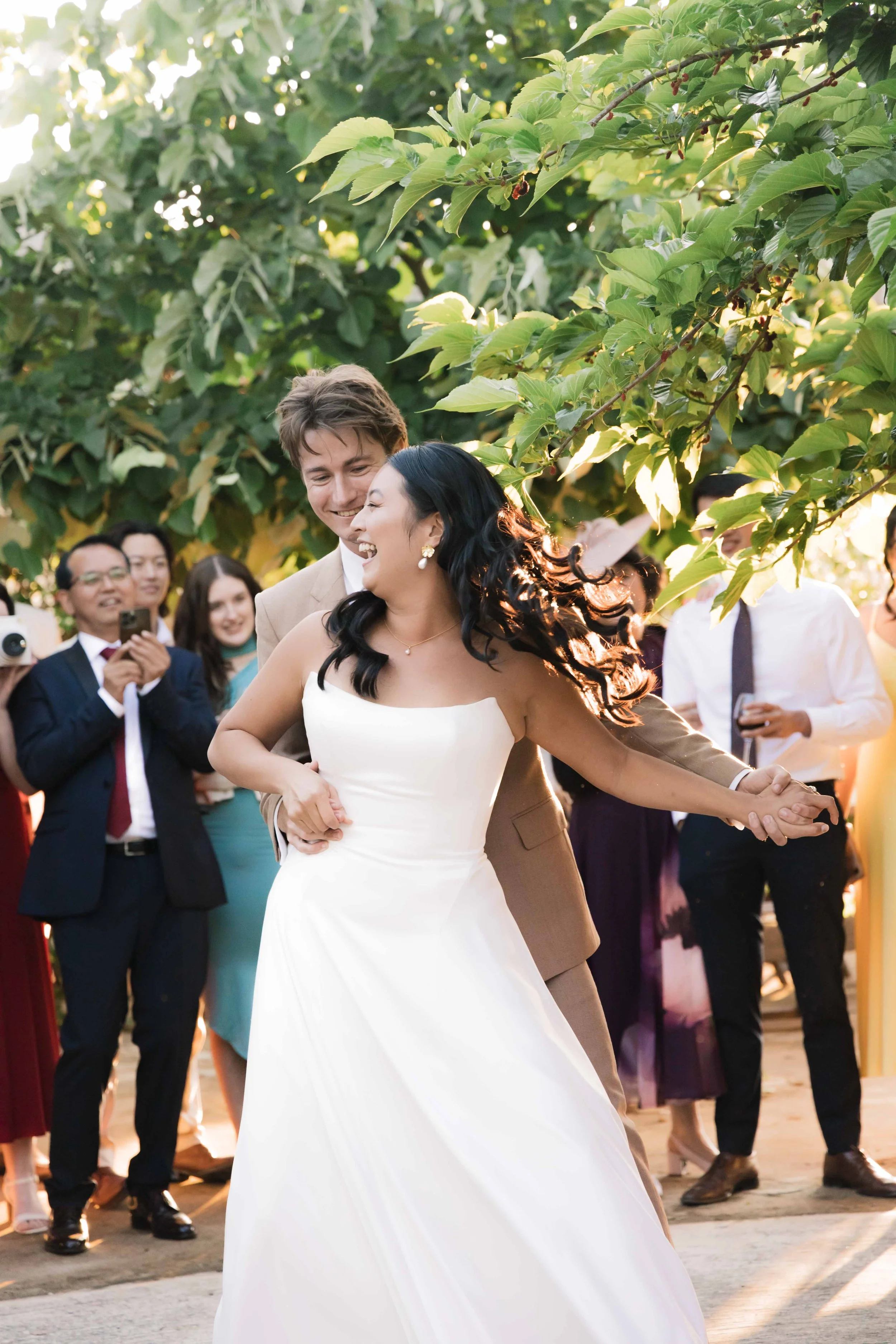 A bride and groom are dancing at their wedding reception outdoors during sunset, surrounded by smiling guests.