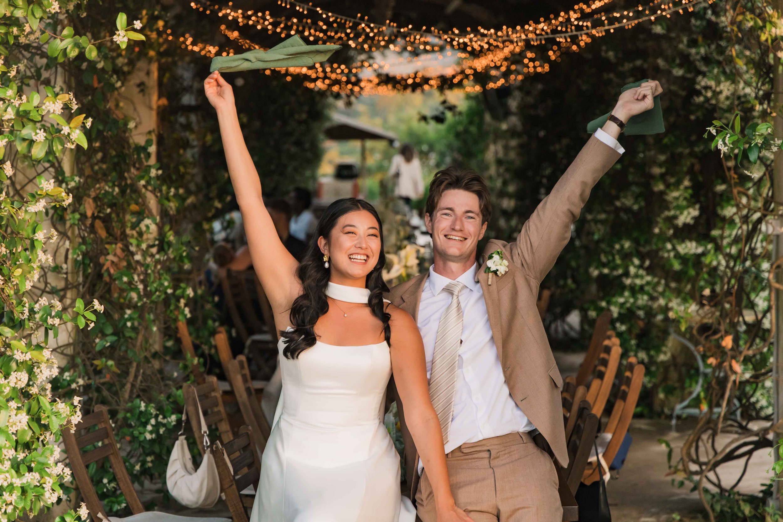 A happy couple, the bride and groom, celebrating at their wedding reception in an outdoor garden area decorated with string lights and greenery, with chairs and guests in the background.
