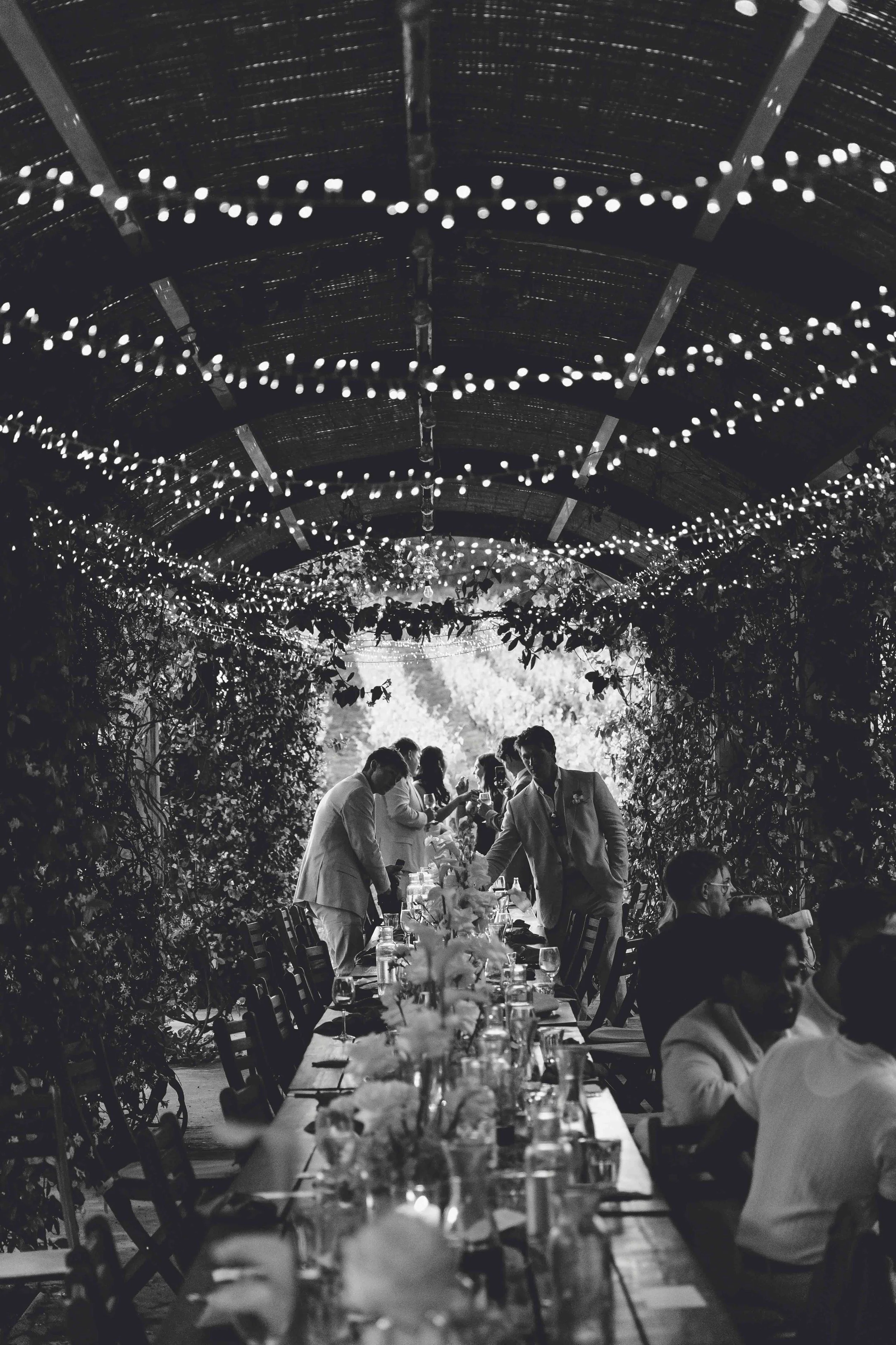 A black-and-white photo of a wedding reception with guests seated at a long table decorated with flowers, under a ceiling decorated with string lights, with some guests standing and conversing.