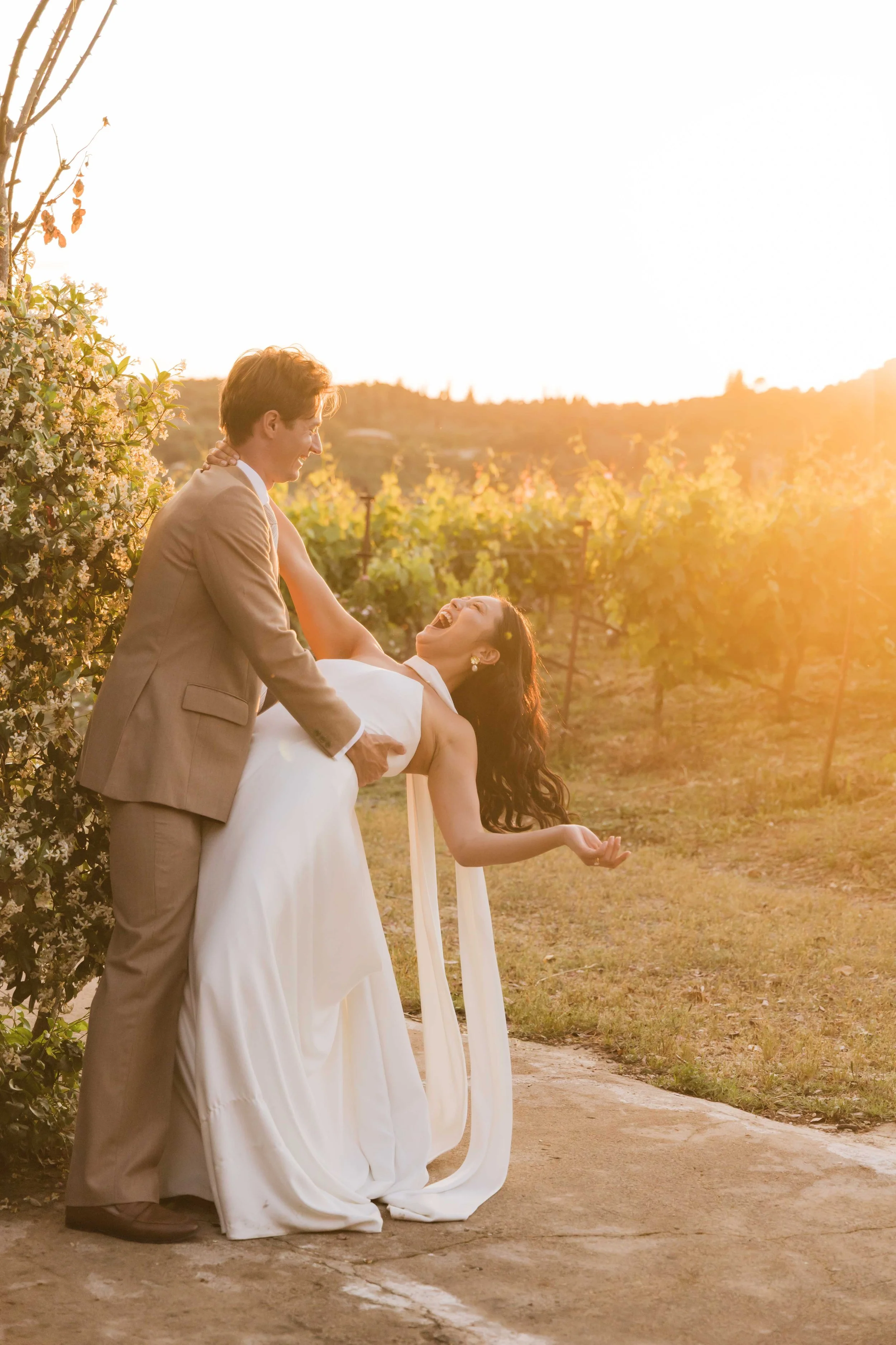 A man and woman in wedding attire sharing a joyful dance outdoors at sunset in a vineyard.