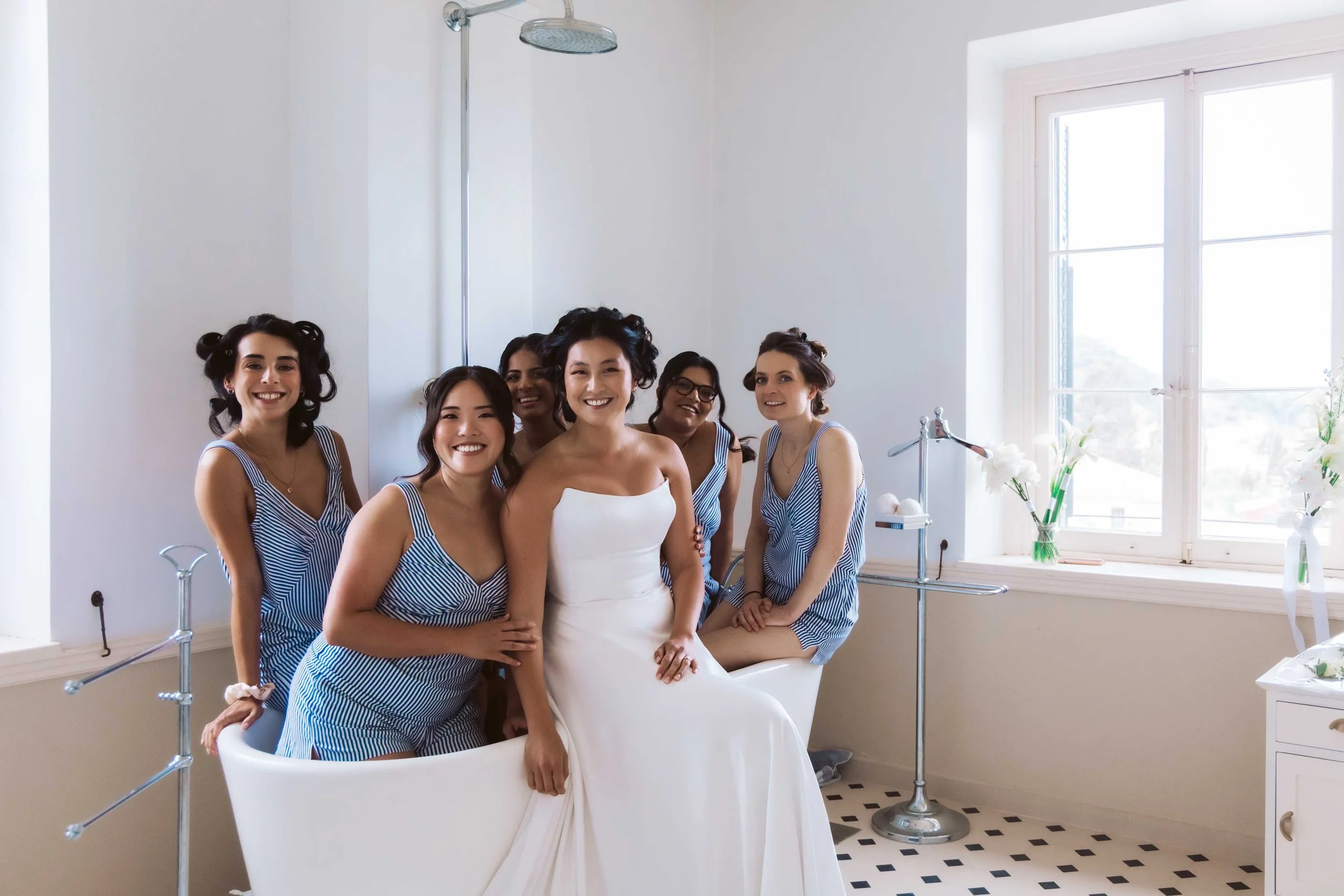 A bride and five bridesmaids pose together in a bright room with large windows, smiling at the camera.