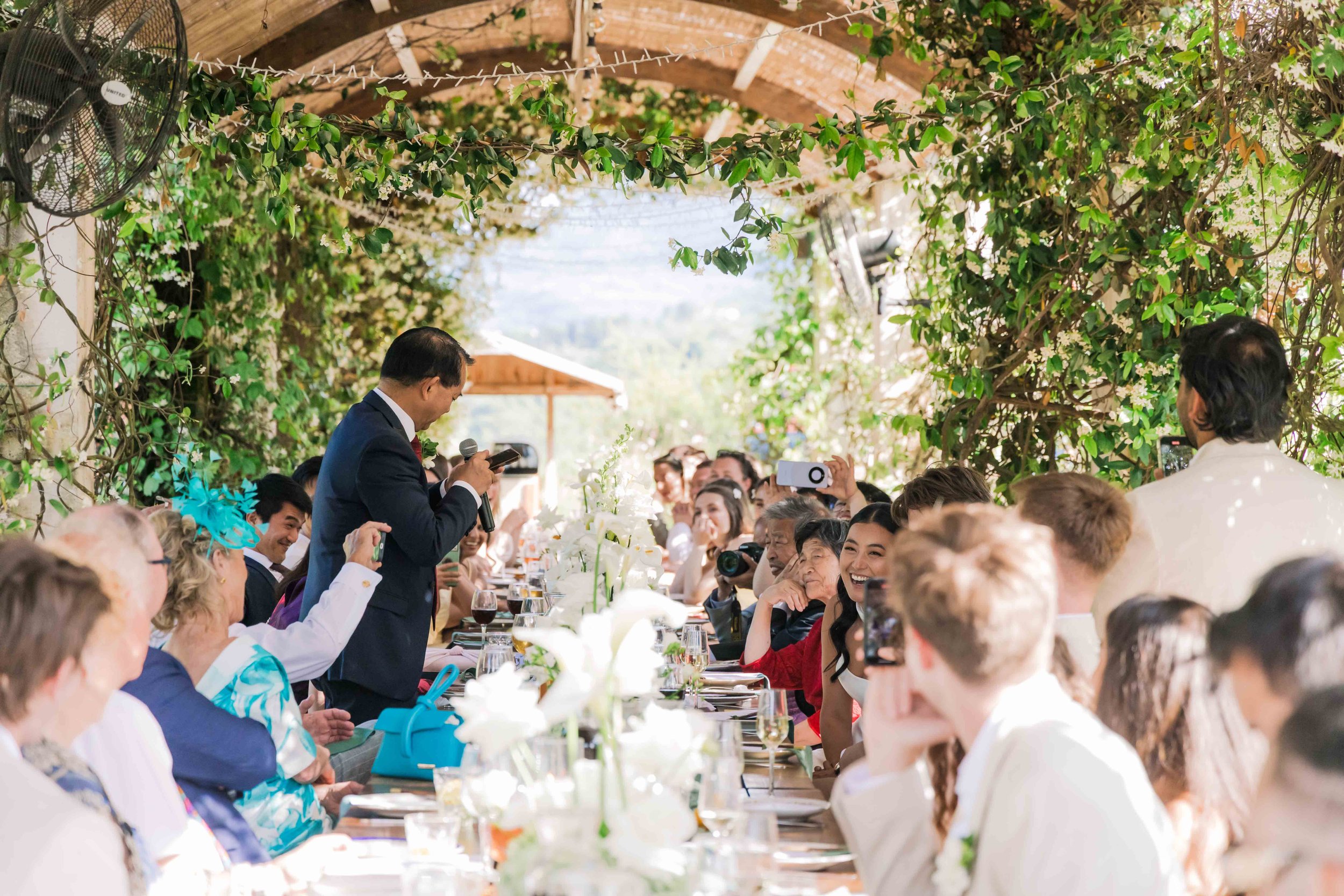 A man in a suit giving a speech at a wedding reception, audience members smiling and taking photos, decorated with greenery, flowers, and string lights.