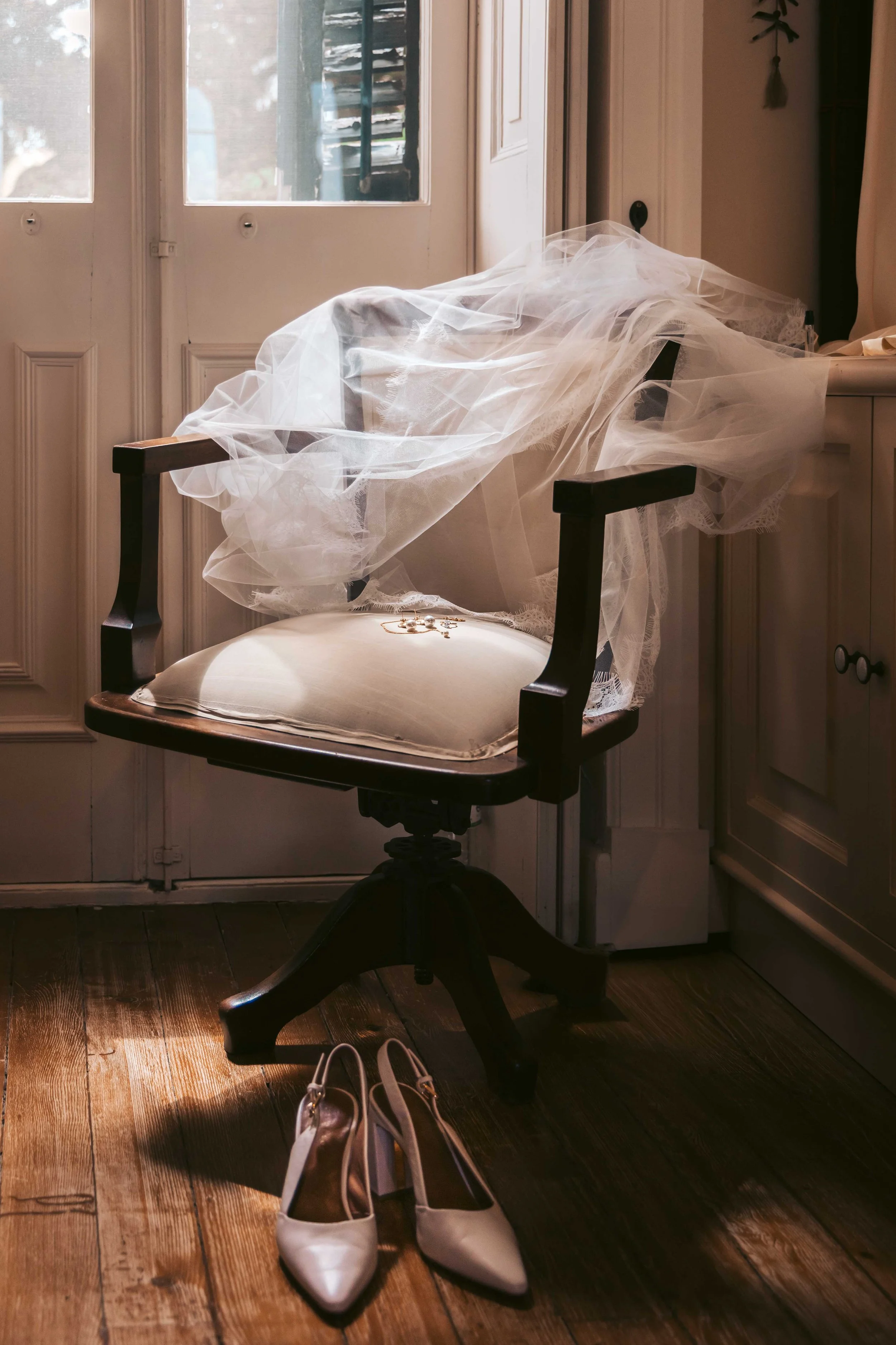An antique wooden chair with a white cushion, covered with bridal veil and jewelry, next to a pair of white high-heeled shoes on a wooden floor by a door with glass panels.