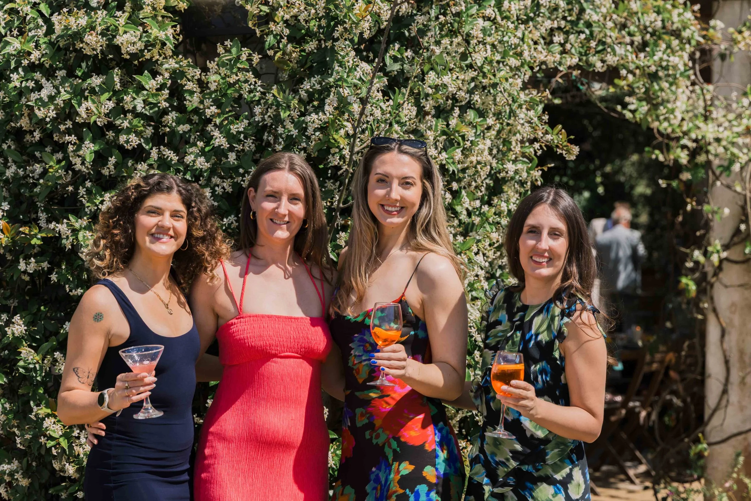 Four women smiling and holding glasses with orange-colored drinks in an outdoor setting with green foliage and white flowers in the background.