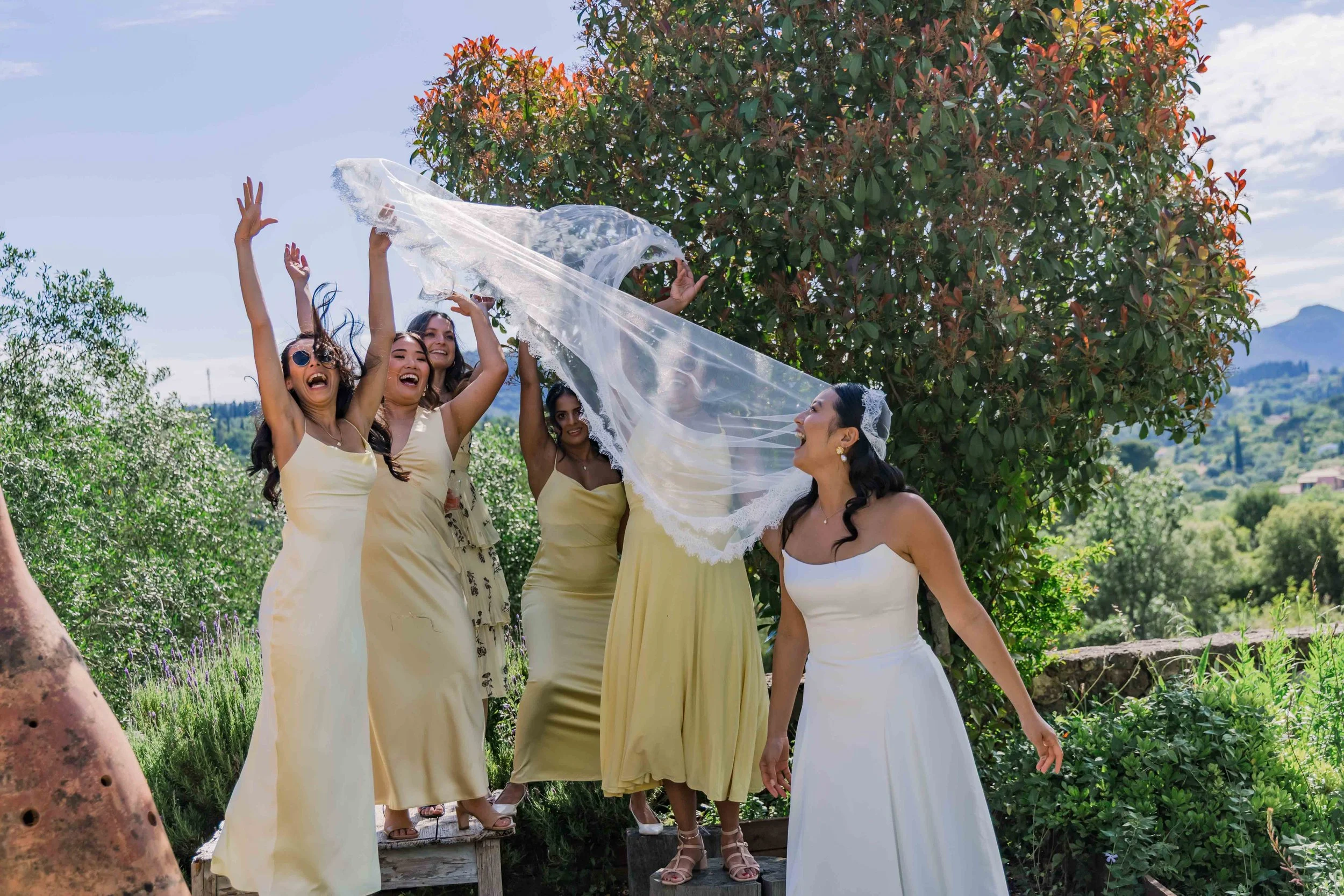 Bride and bridesmaids celebrating outdoors on a sunny day, with one bridesmaid tossing a veil towards the bride, surrounded by greenery and mountains in the background.