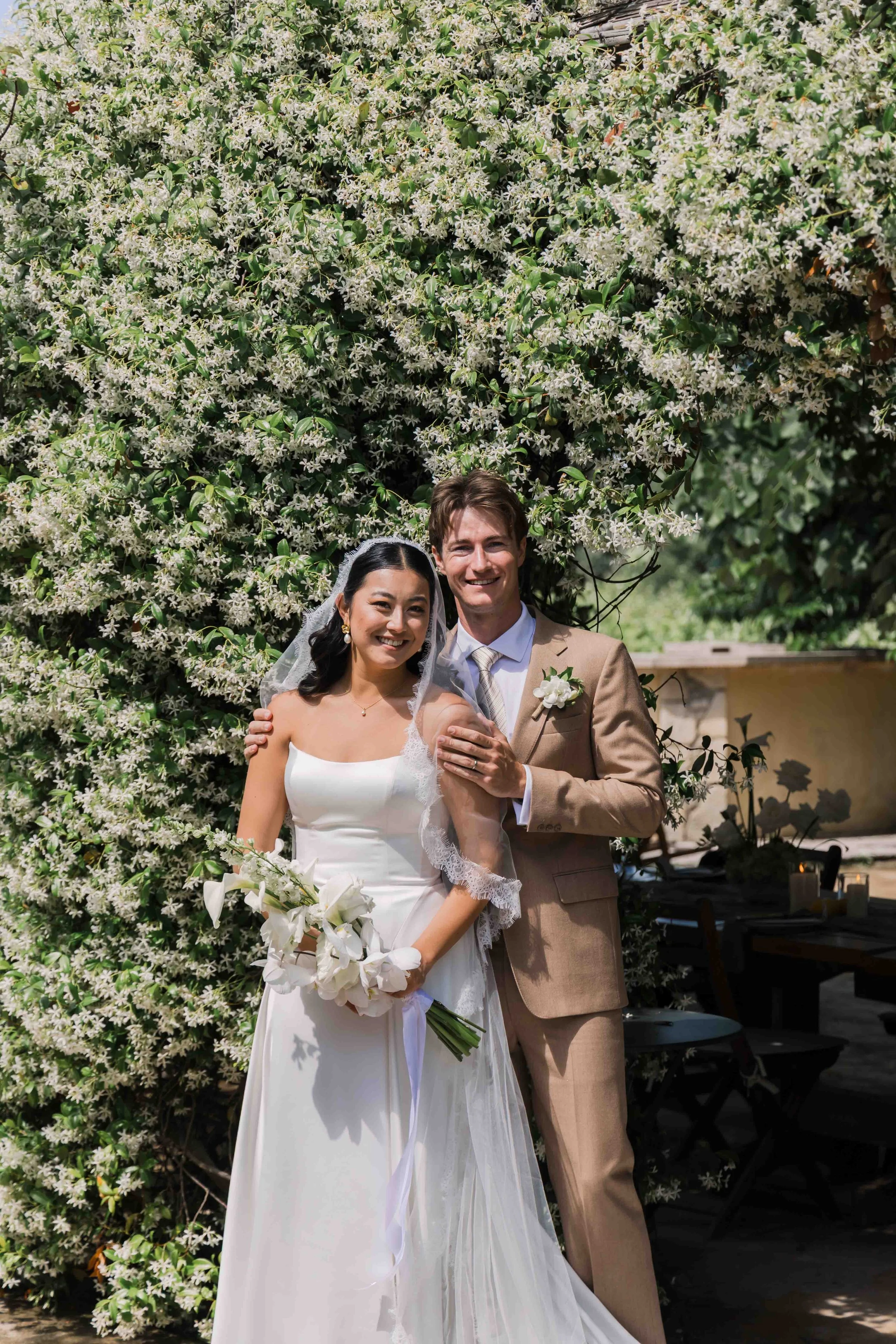A smiling bride and groom standing close together outdoors under a flowering bush, with the bride holding a bouquet of white flowers.