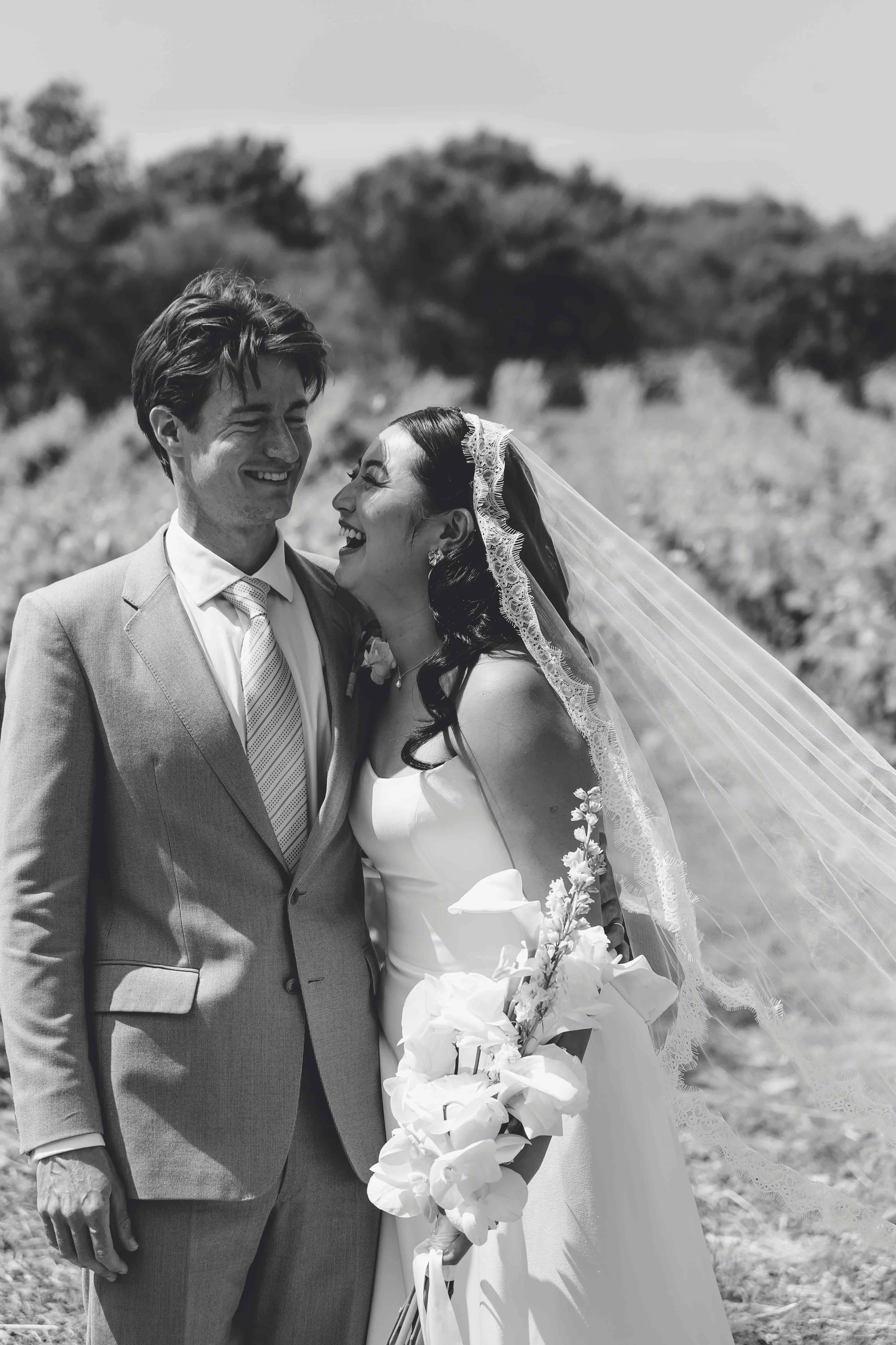 Black and white photograph of a newlywed couple smiling and looking at each other outdoors, with trees in the background. The bride is wearing a lace veil and holding a bouquet of white flowers, while the groom is dressed in a light suit with a patte