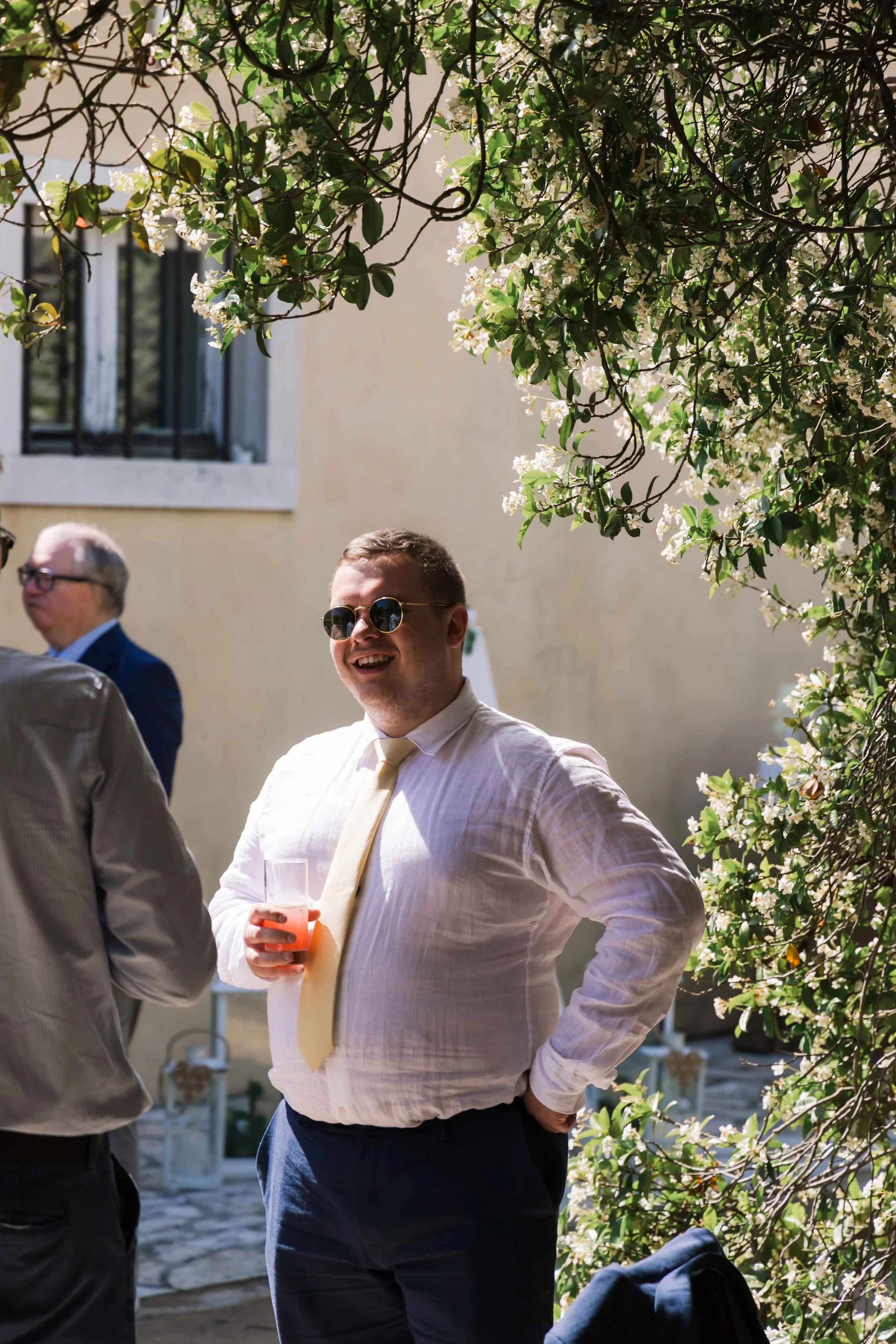 A man wearing sunglasses, a light-colored shirt, and a light yellow tie is smiling and holding a glass of pink beverage at an outdoor event. There are other people in the background, and flowering greenery surrounds the scene.