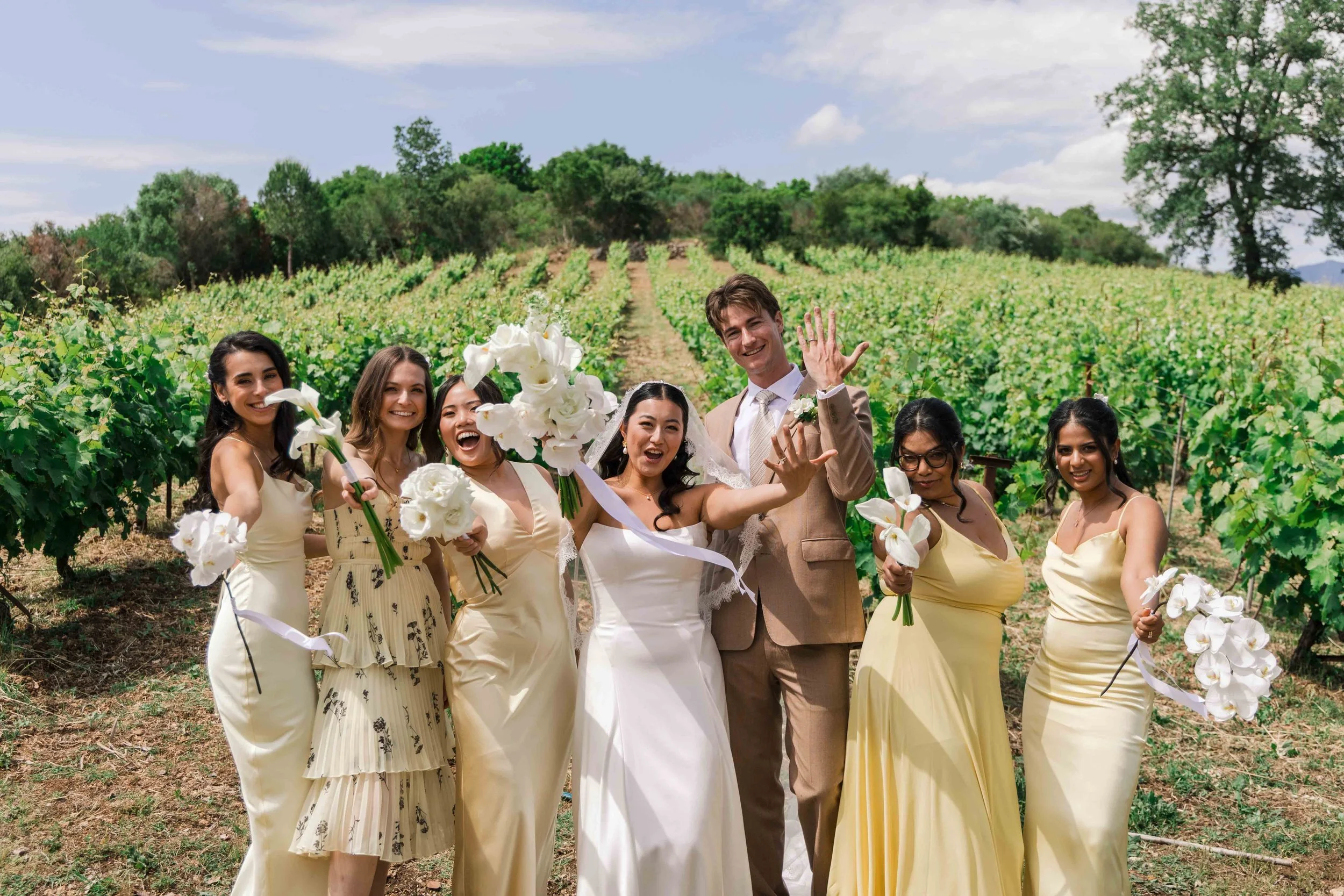 Wedding party of seven people on a vineyard, including the bride in a white dress and groom in a tan suit, surrounded by bridesmaids holding white bouquets, celebrating outdoors on a sunny day under partly cloudy sky.