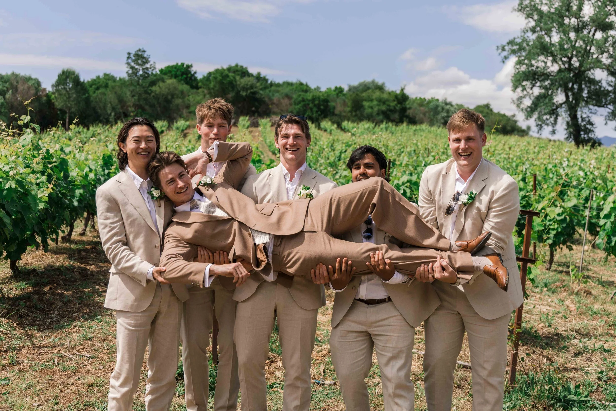 Group of seven men in beige suits, with one man lying across the others' arms, in a vineyard during daytime, smiling and posing for the photo.