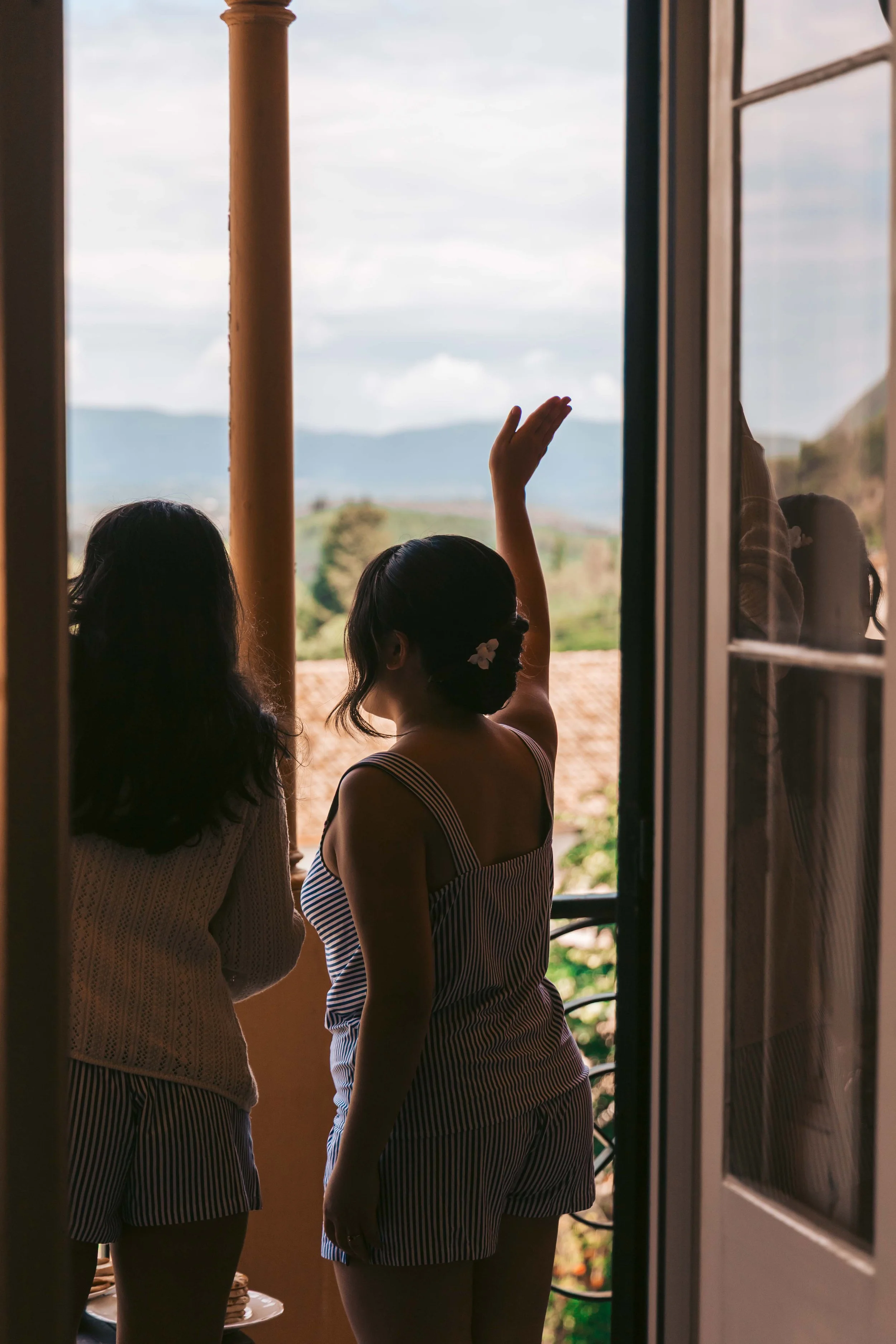 Two women standing on a balcony, looking out at a scenic landscape with mountains, trees, and a cloudy sky, one woman raising her hand.