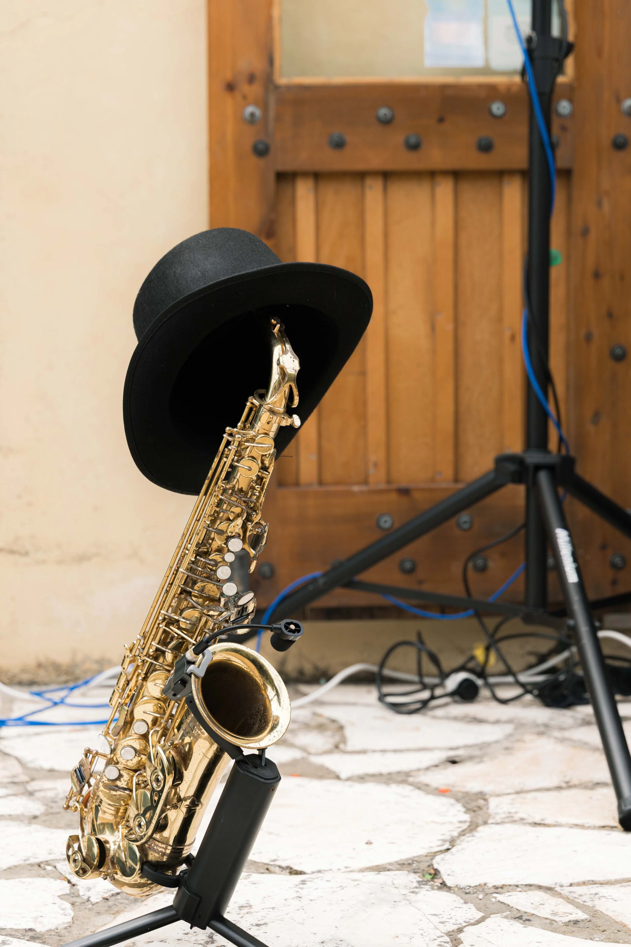 A saxophone with a black fedora hat on its neck strap, set on a stand outside near a stone pathway, with a wooden door and some wires in the background.