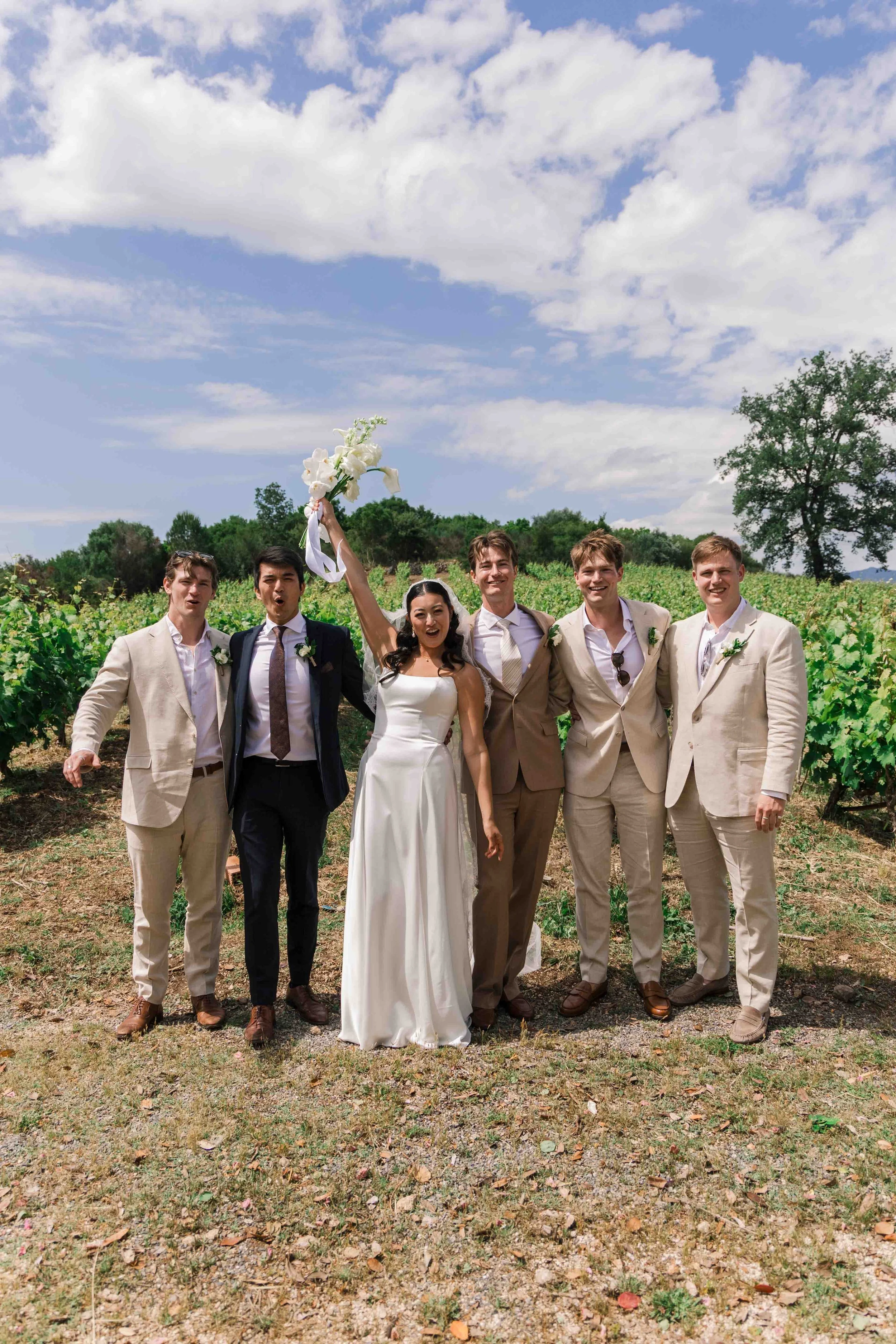 A bride and five groomsmen standing outdoors in a vineyard, celebrating, with the bride holding a bouquet in the air.