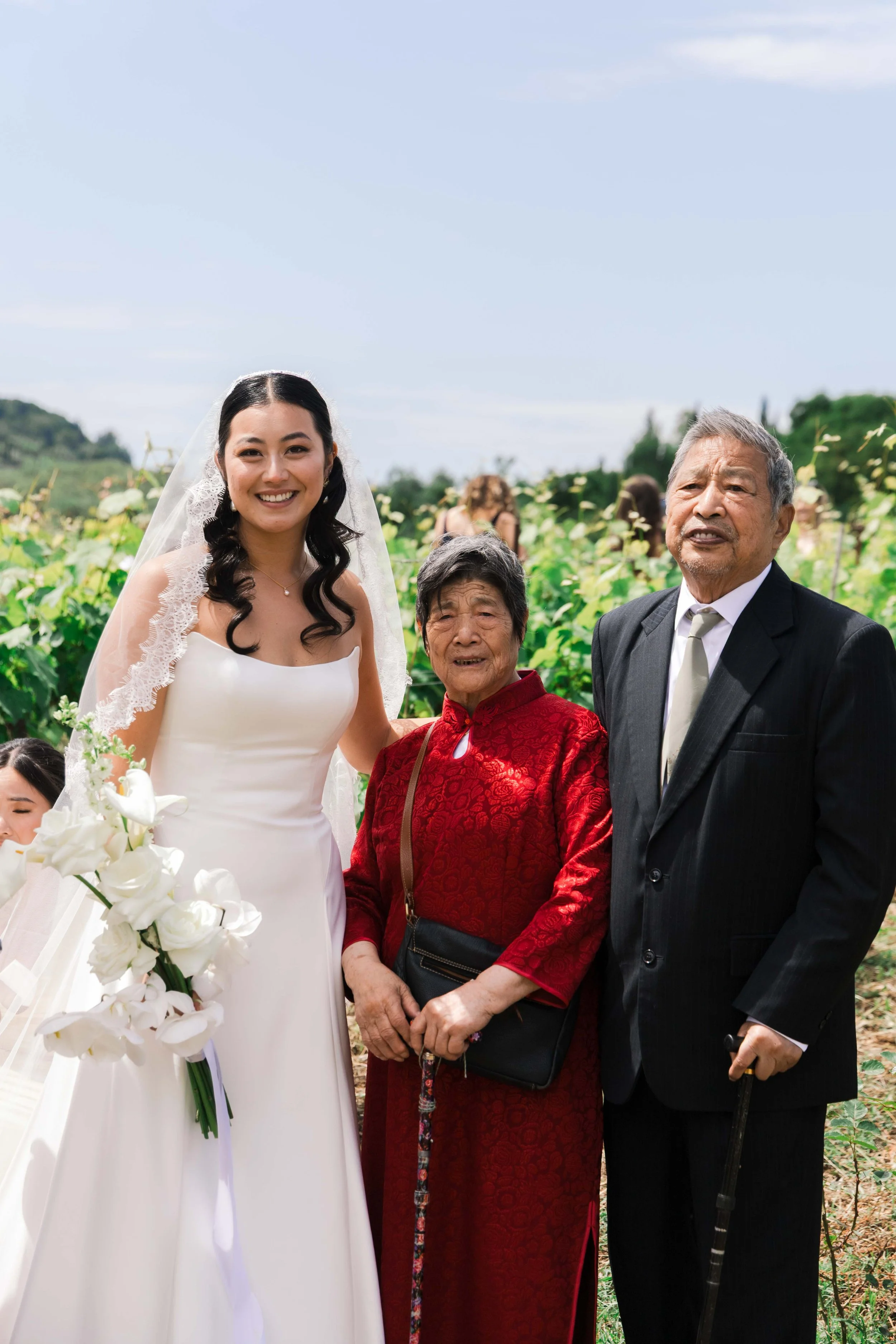 A bride in a white wedding dress and veil standing outdoors with her elderly parents, a woman in a red dress and a man in a black suit, in a lush green field on a sunny day.