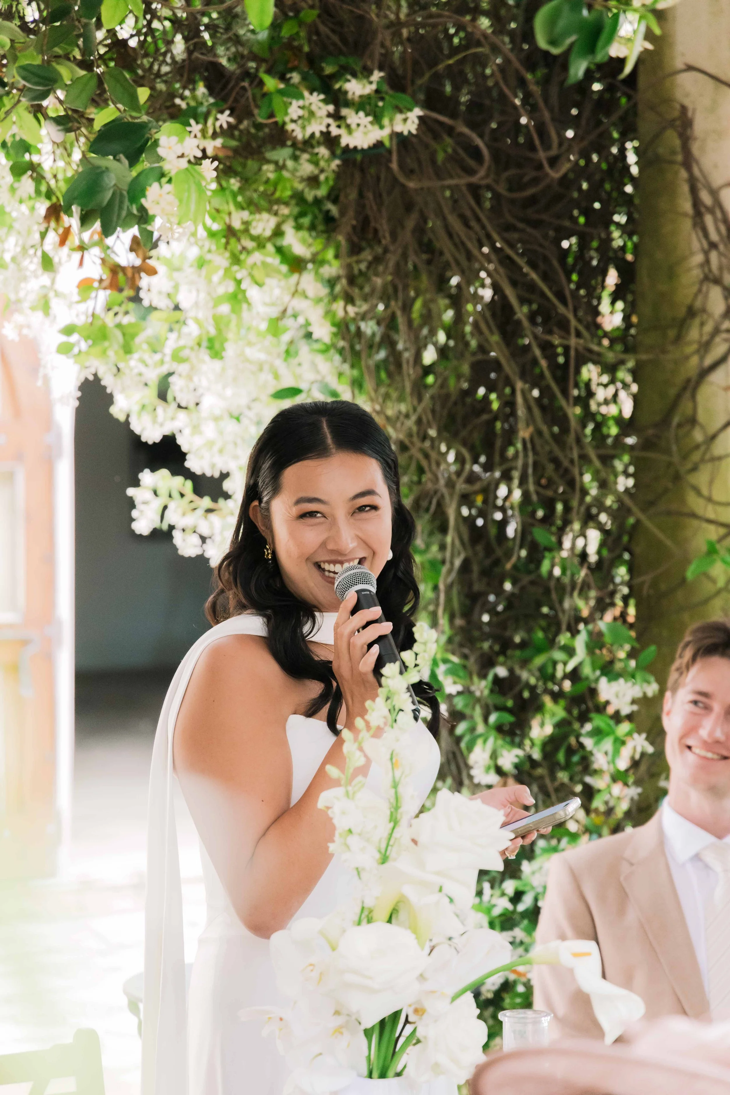 A woman in a white dress smiles and speaks into a microphone at a wedding reception, with a man in a beige suit sitting nearby. They are outdoors under a floral arch with white flowers and green foliage.