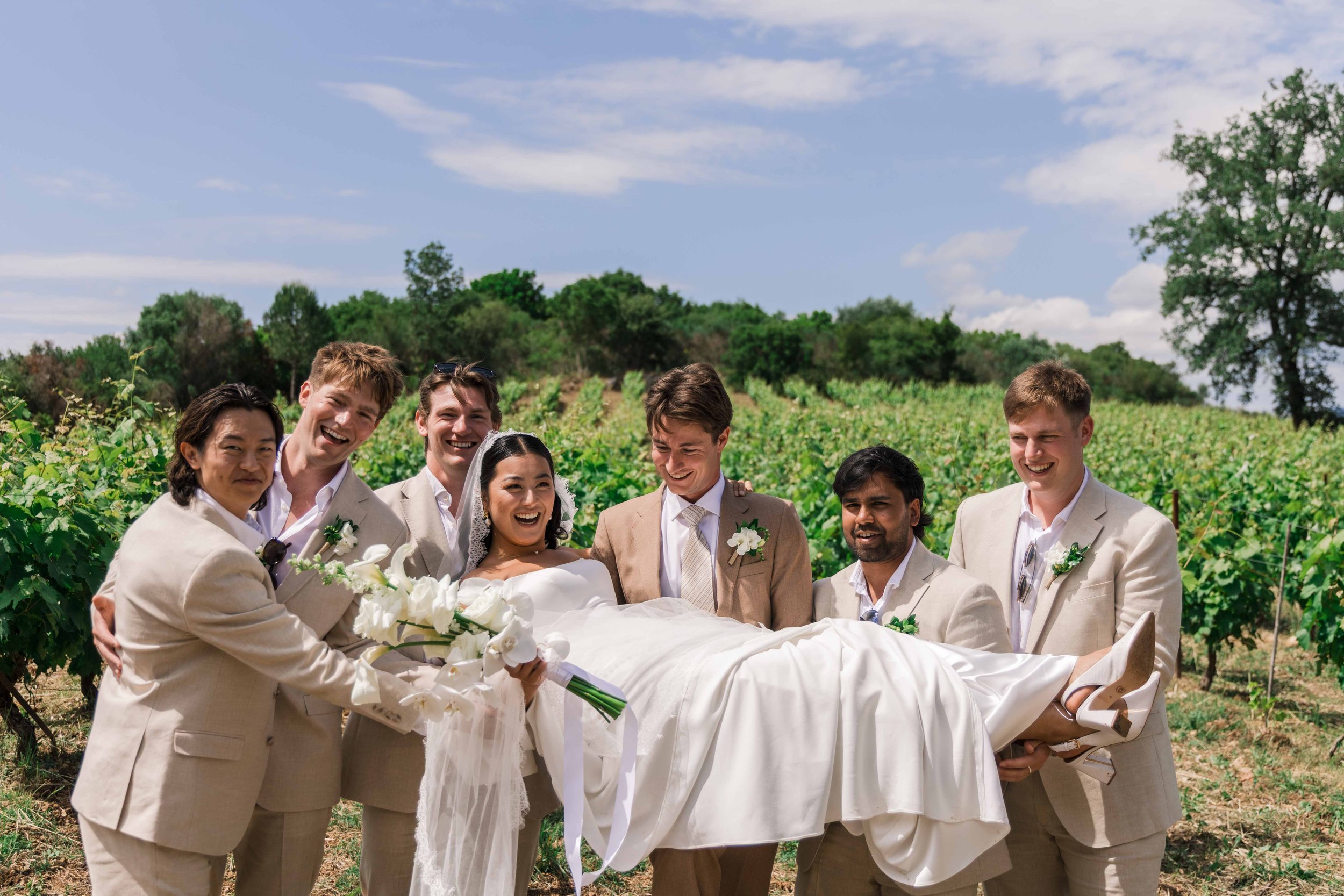 A bride in a white wedding dress being carried by several groomsmen in light-colored suits in a vineyard, all smiling and joyful on a sunny day.