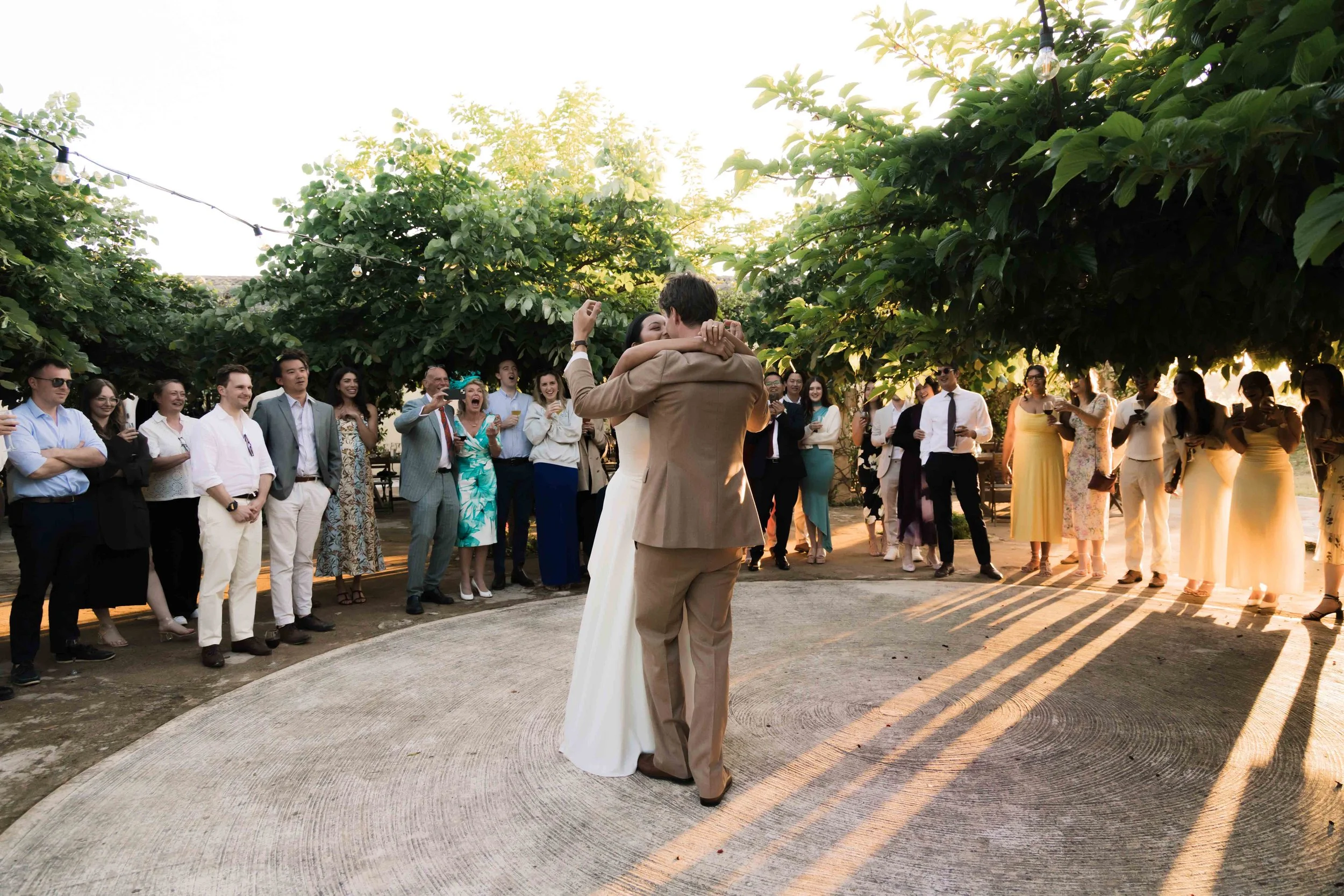 A bride and groom are dancing outdoors at their wedding reception, surrounded by guests under large trees with hanging string lights, during sunset.
