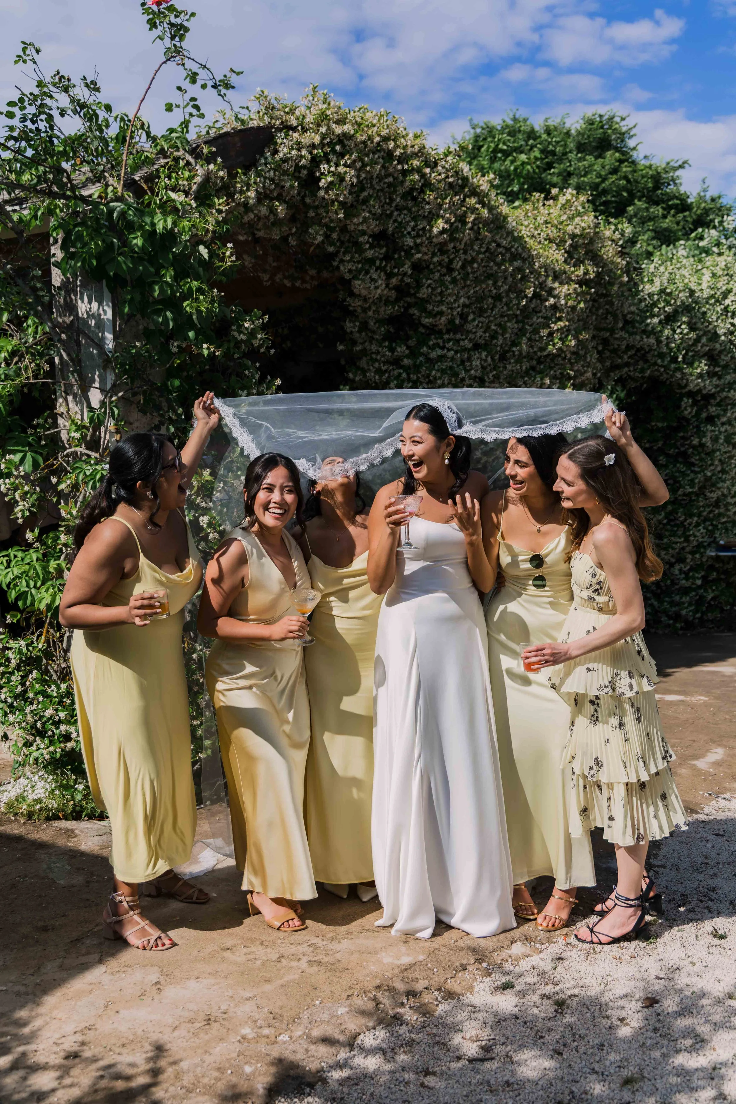 Group of women in dresses celebrating outdoors, with one woman in a white wedding dress, holding a glass, surrounded by friends sharing a joyful moment.