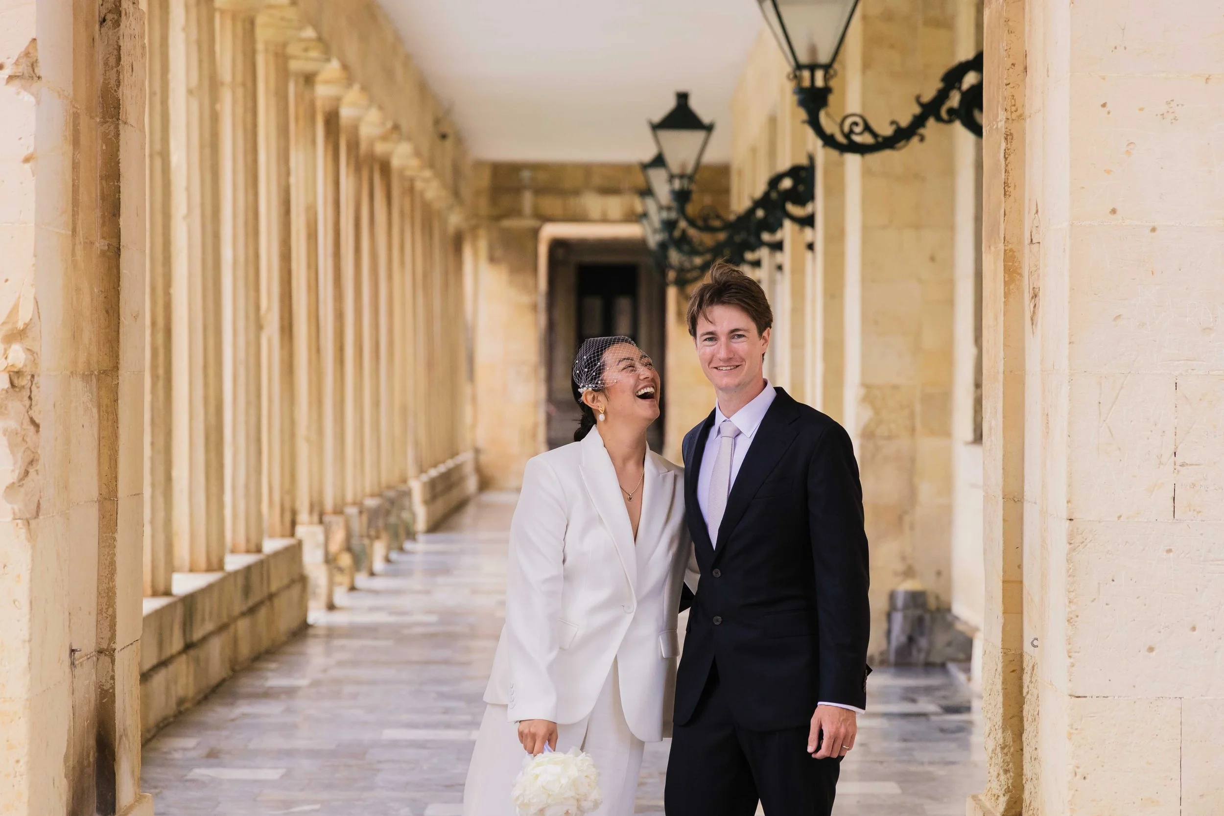 A woman and a man, both dressed in wedding attire, standing in a stone corridor with lantern-style lights overhead. The woman is laughing and holding a bouquet, the man is smiling.