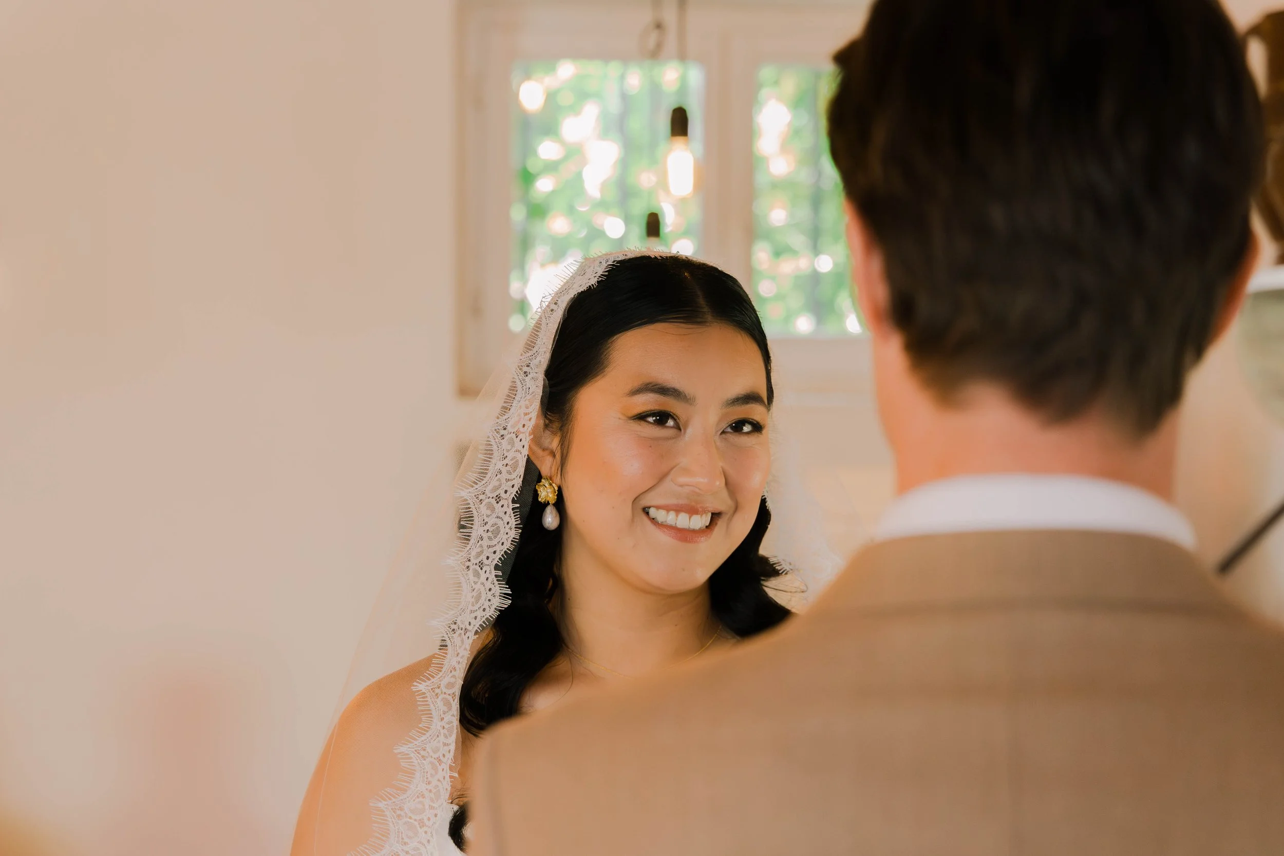 Bride and groom smiling at each other indoors with soft lighting and windows in the background.