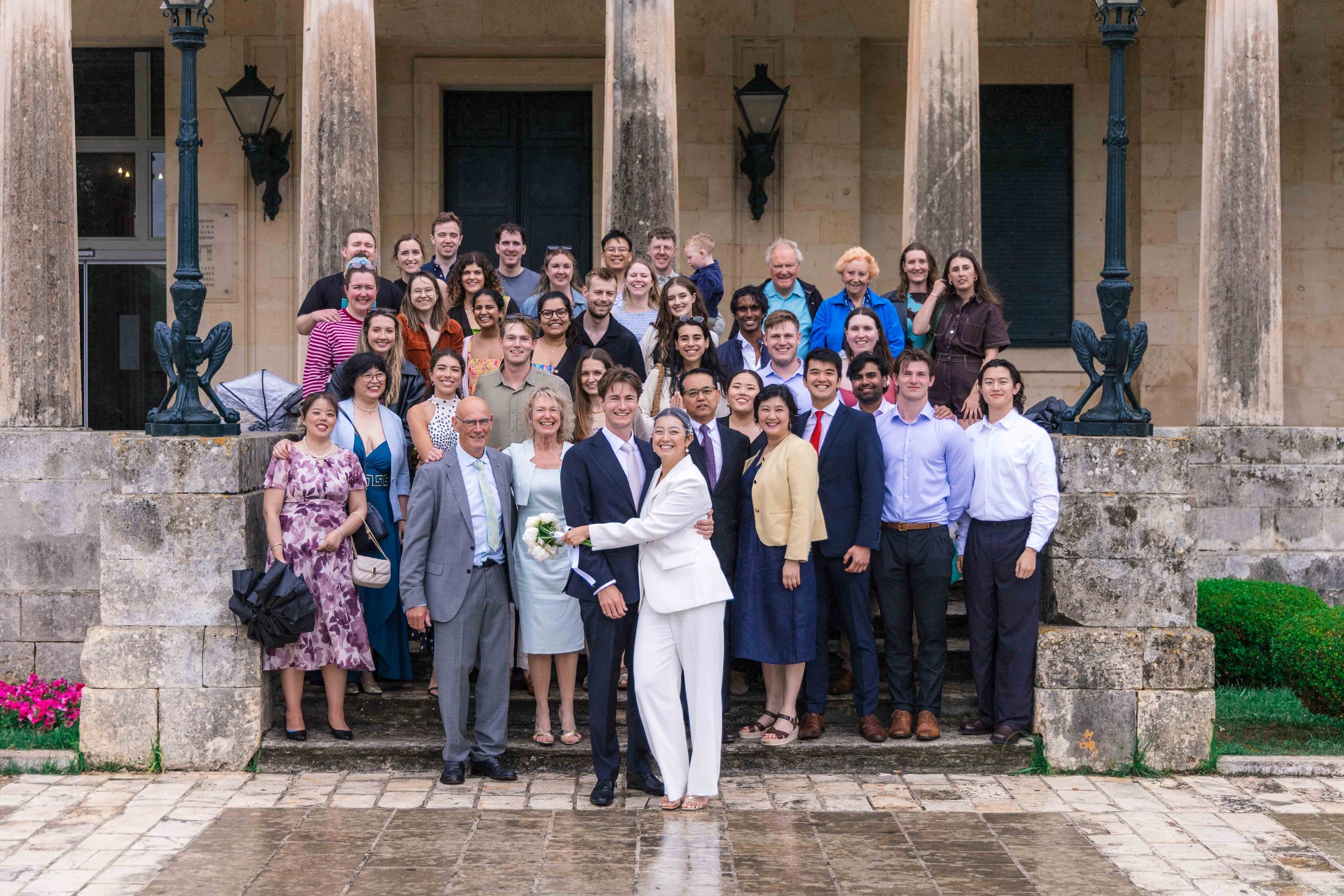 A large group of people gathered on the steps outside a historic building with stone columns, celebrating a wedding. The bride and groom are at the center, smiling and holding flowers. The group includes men and women of various ages dressed in forma