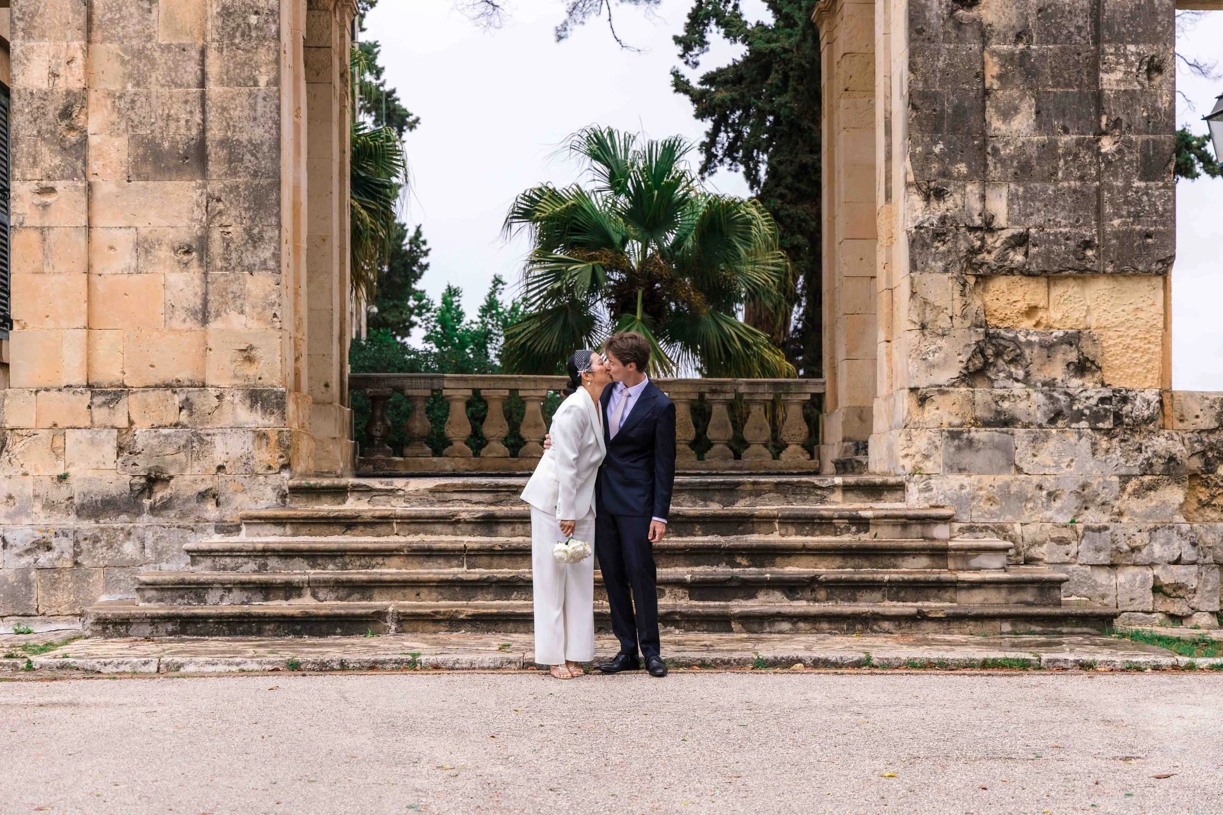 A newlywed couple sharing a kiss on old stone steps in front of a historic building with ornate stone columns and greenery in the background.
