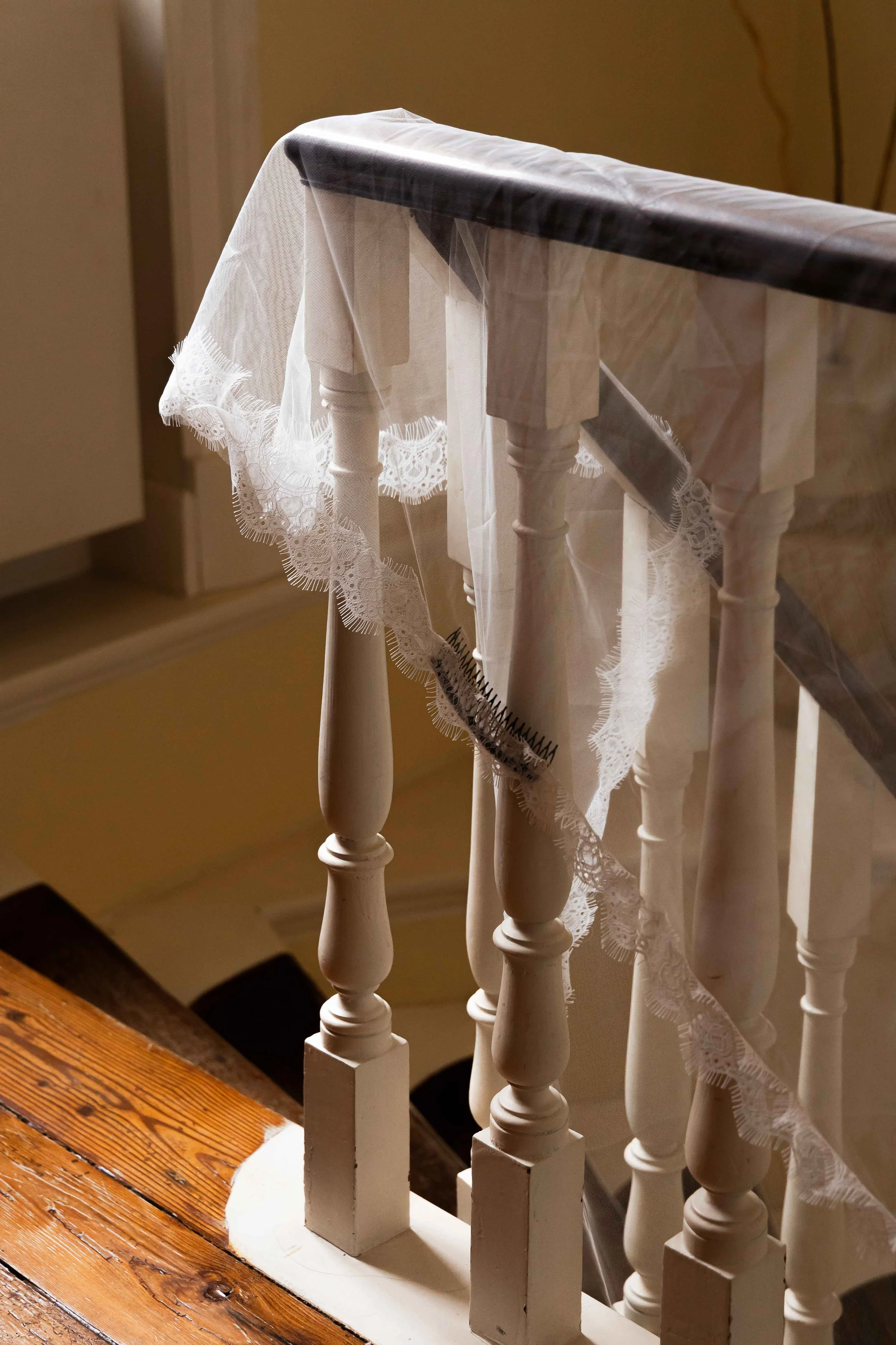A white lace curtain draped over a set of white wooden balusters on a staircase, with sunlight shining through.