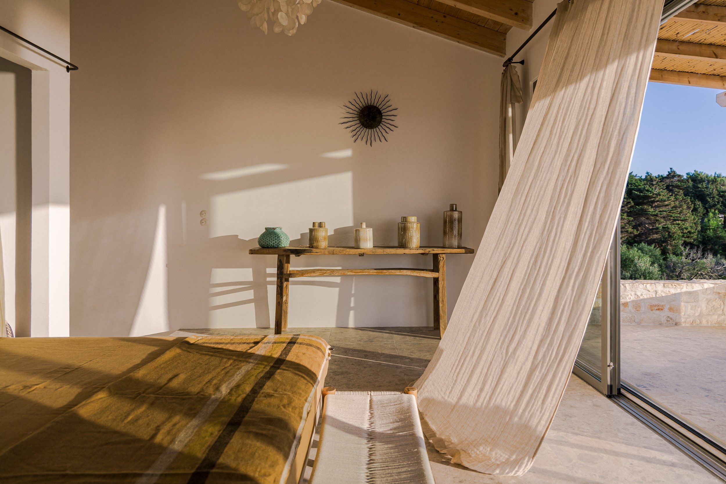 Sunlit interior of a room with a beige curtain partially open, revealing a view of trees outside. A wooden table against the wall holds five decorative vases in neutral tones. Shadows cast by sunlight create a warm atmosphere.