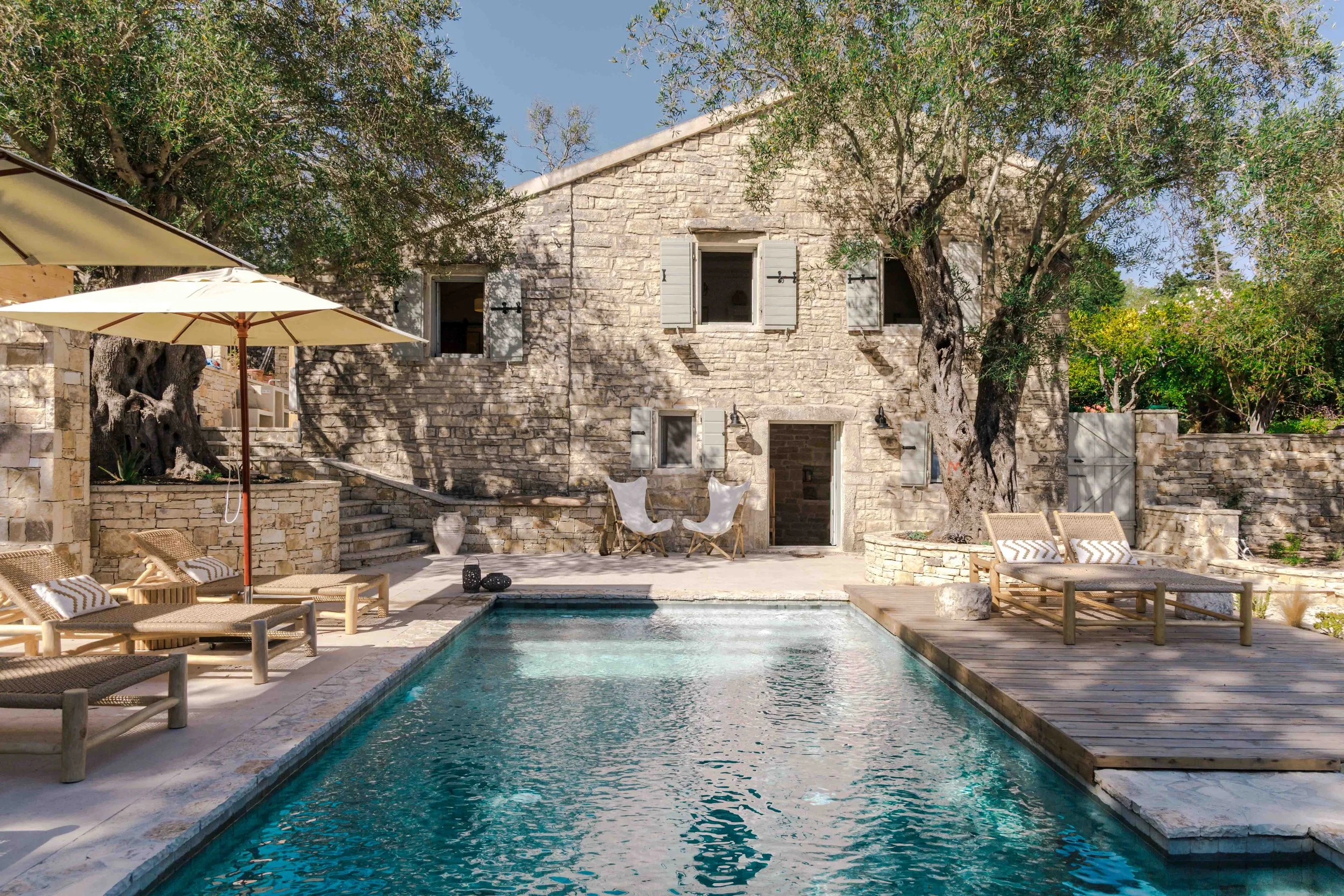 Stone house with open windows, large trees, outdoor pool, sun loungers, umbrellas, and patio furniture.