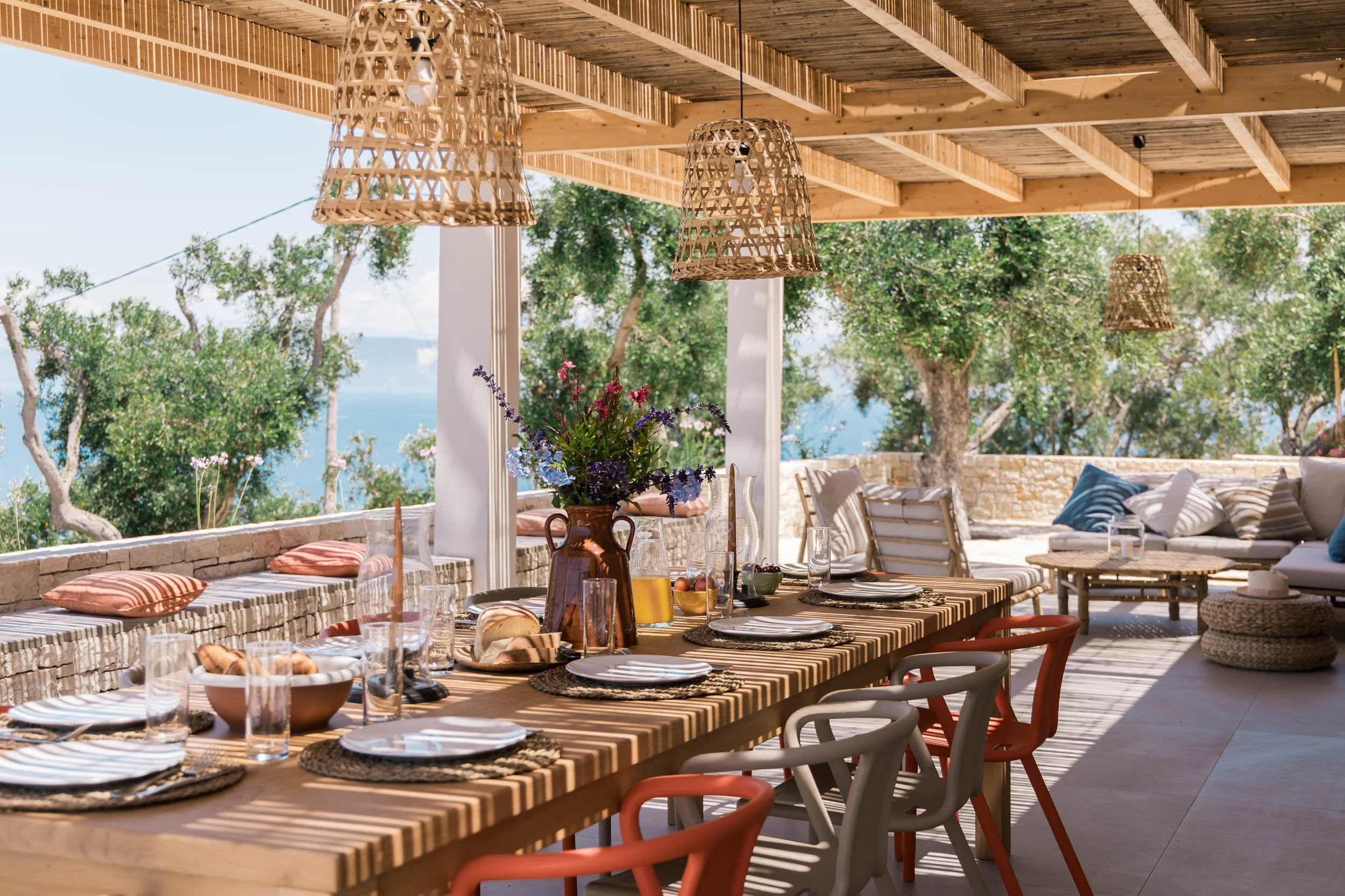 Outdoor dining area with a wooden table set with plates, glasses, bread, and a flower vase, under a wooden ceiling with woven pendant lights, overlooking trees and a body of water in the background.