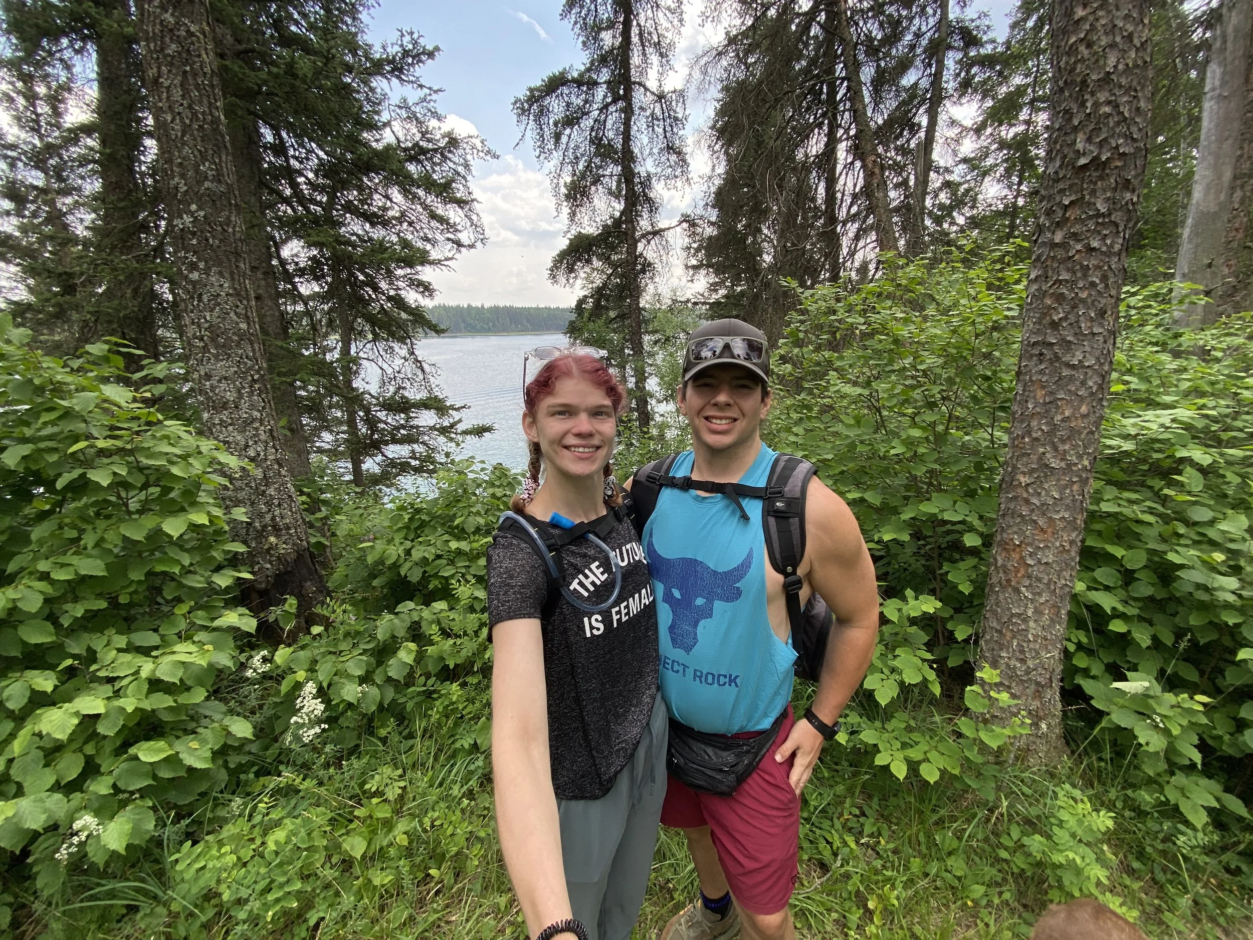 Two smiling young hikers in outdoor gear pose for a selfie in a lush forest with a lake in the background.