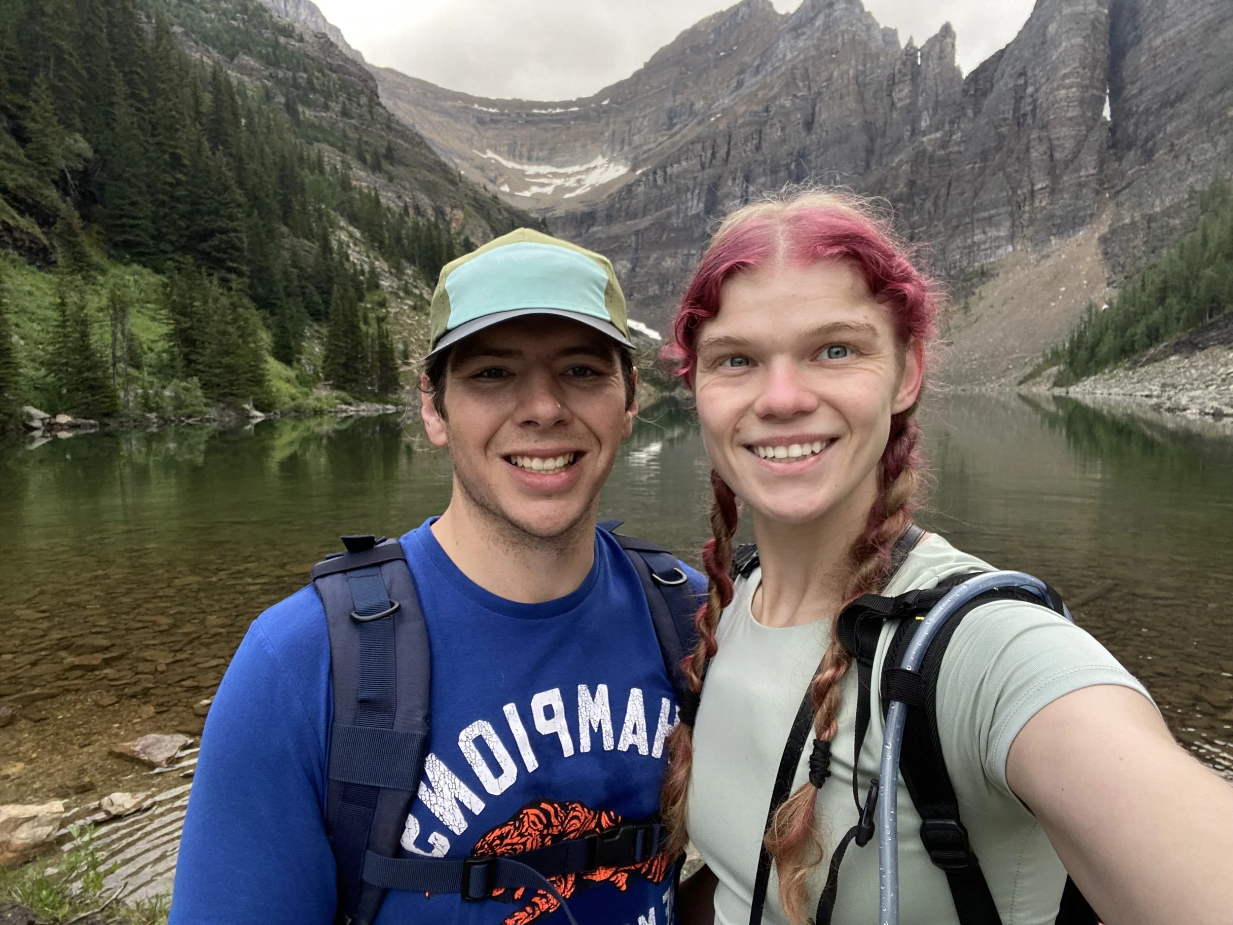 Two smiling hikers, a young man wearing a blue T-shirt and a young woman with pink hair in braids wearing a light green shirt, posing for a selfie near a lake in a mountain landscape with steep rocky cliffs and dense green trees.
