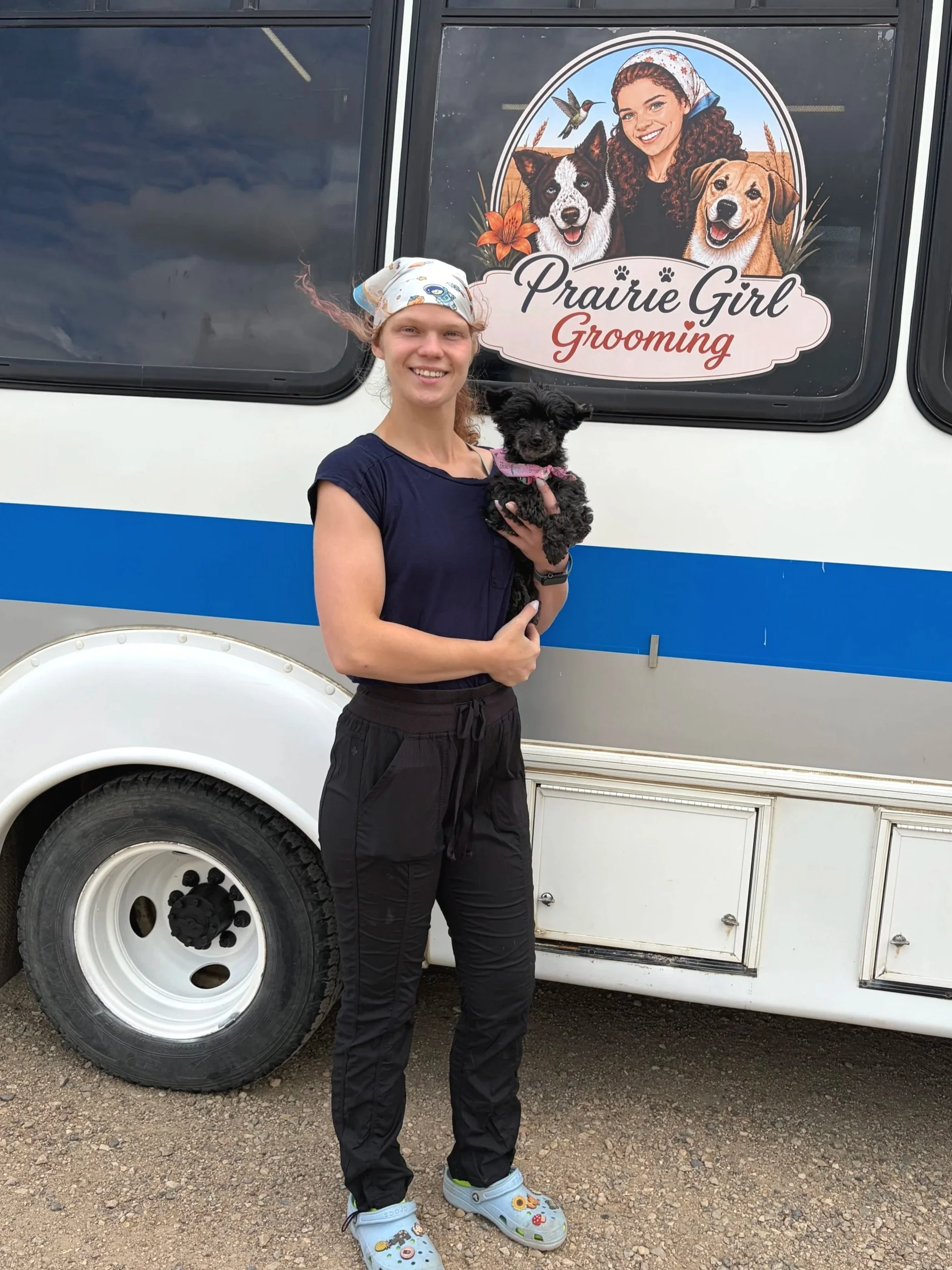 A young woman with curly hair smiling while holding a small black dog with a pink bandana. They are standing outside in front of a trailer with a large sign that says "Prairie Girl Grooming" and features illustrations of a woman with two dogs and a hummingbird.