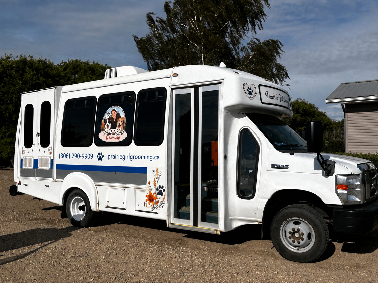 A white grooming van with decals for Prairie Girl Grooming, featuring a woman with a dog and a cat illustration on the side, parked outdoors on dirt ground with trees and a house in the background.