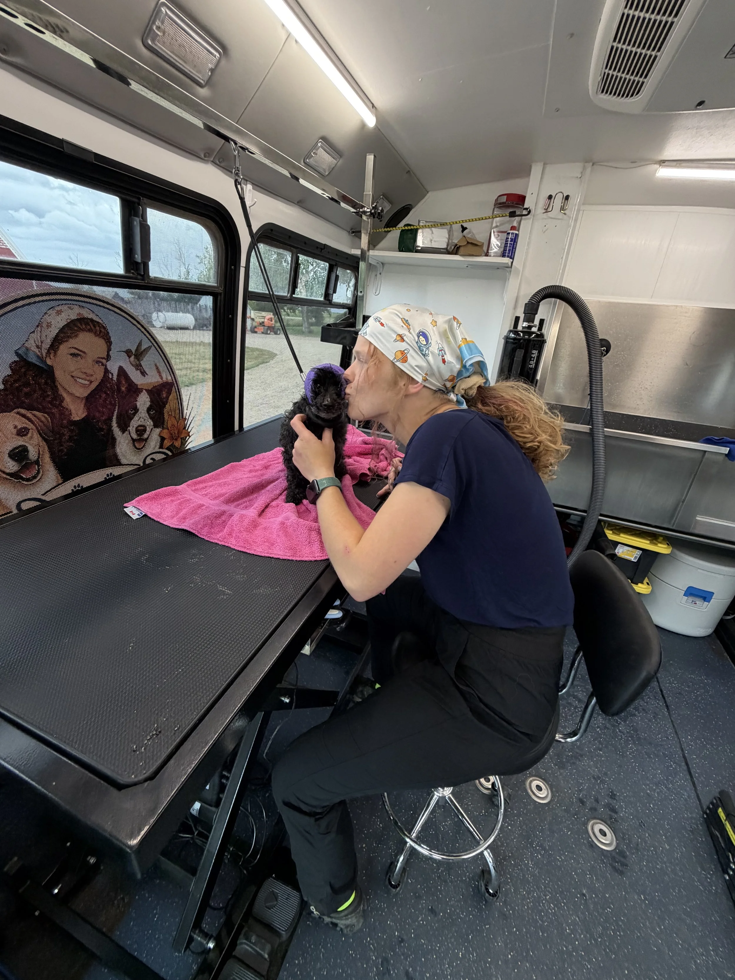 A woman with curly hair, wearing a bandana with space-themed prints, is kissing a small black puppy on a grooming table inside an animal grooming van. The woman is dressed in a navy blue shirt and dark pants, and a pink towel is on the grooming table. The van has a window with a decorative picture of a woman with a dog, and grooming supplies are visible on a shelf above.