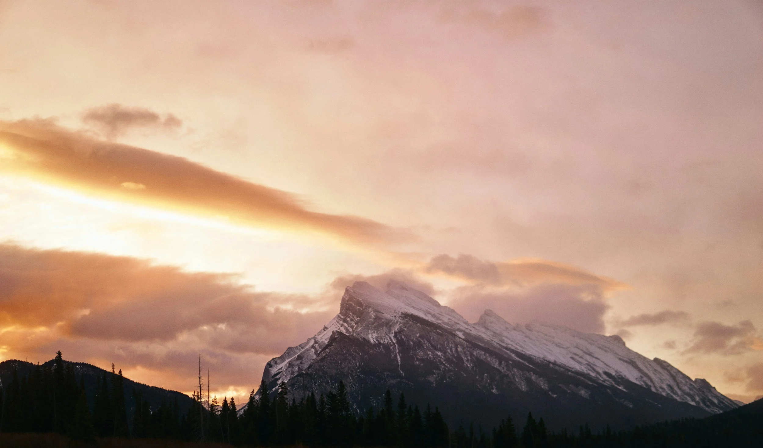 Snow-capped mountain peak during sunset with a colorful sky and clouds in the background.