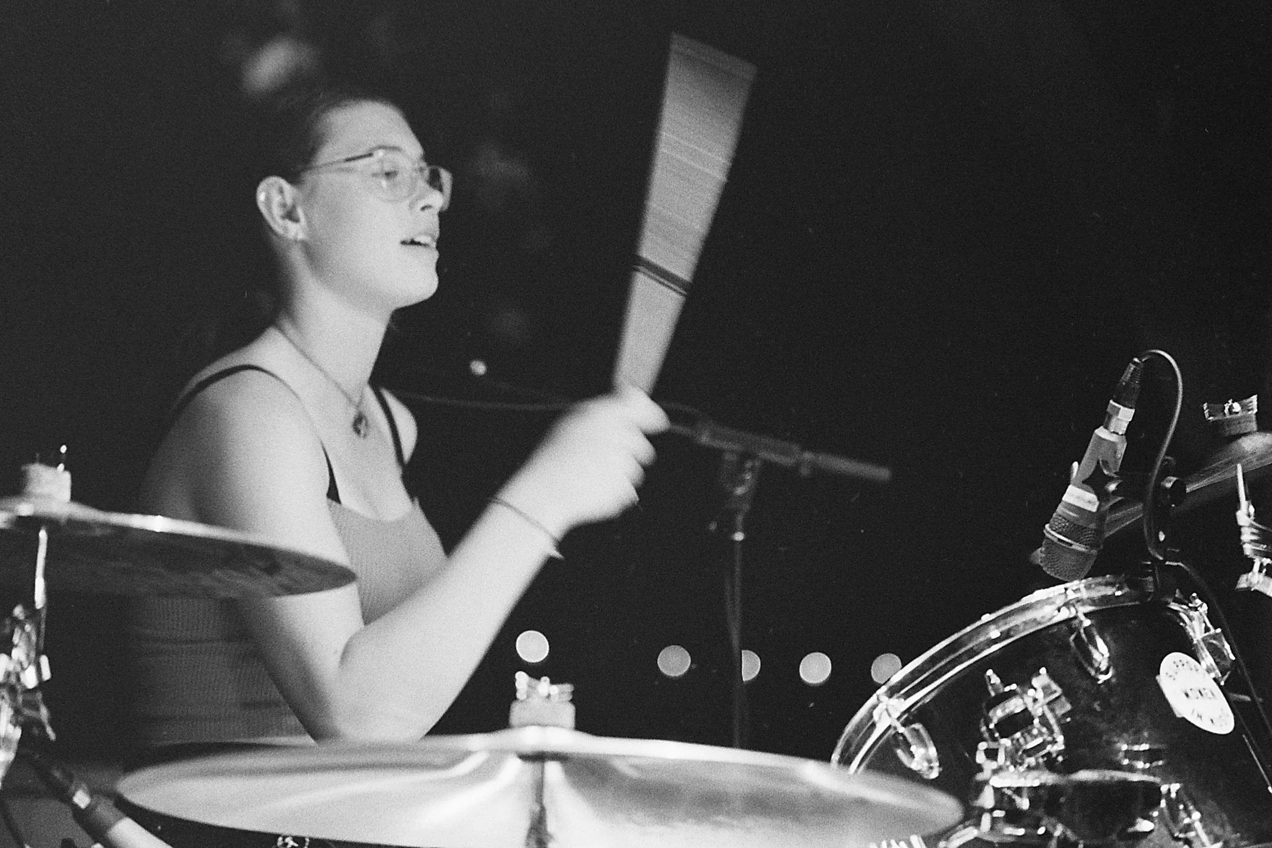 A black and white photo of a woman playing the drums in a recording studio, wearing glasses and a tank top.
