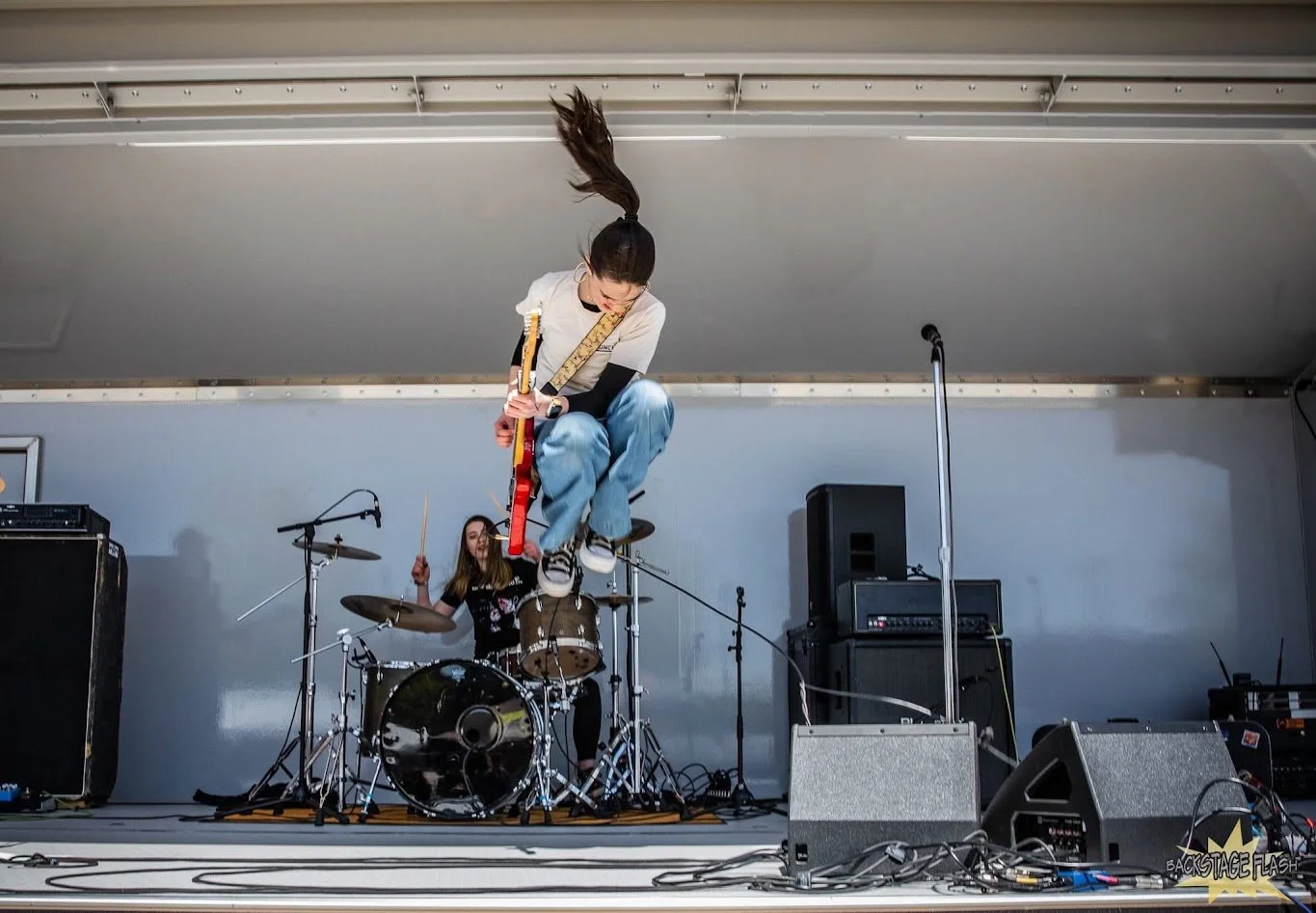 A woman is jumping in mid-air while playing an electric guitar during a live band performance, with a drummer in the background and sound equipment on stage.