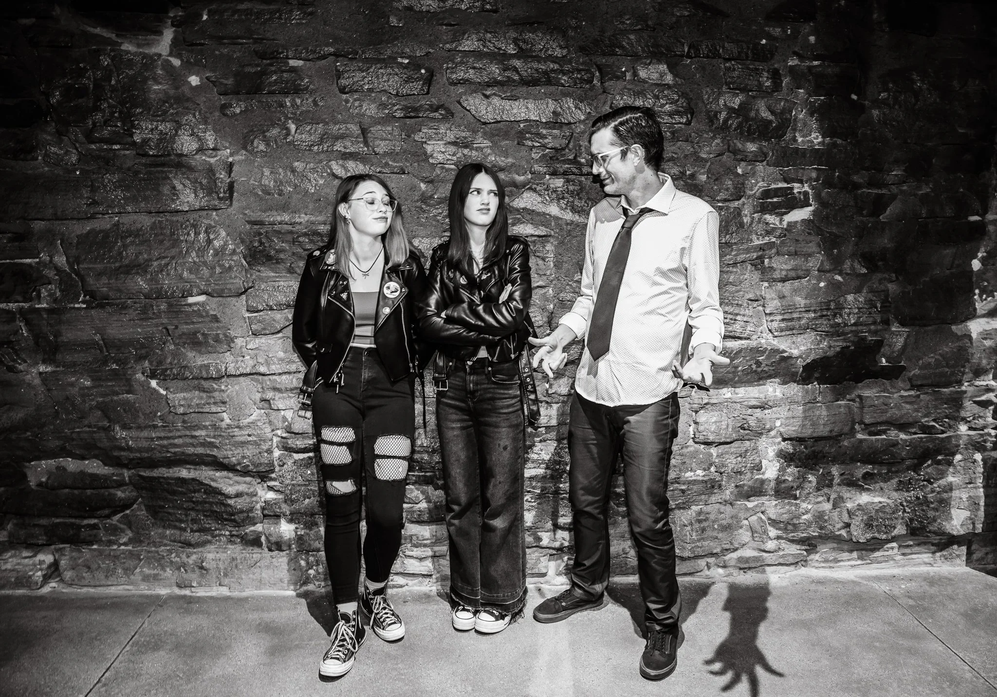 Two young women and a man standing against a stone wall, engaged in conversation. The women are wearing black leather jackets, one with glasses and ripped jeans, the other with straight hair and a black jacket. The man is wearing glasses, a white shi