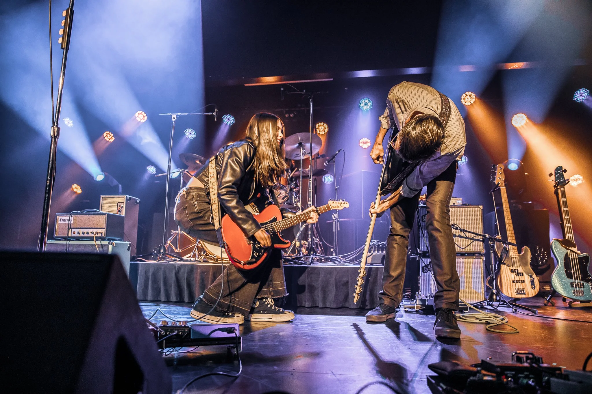 Musicians on stage performing with guitars and microphones, stage lights shining above them.