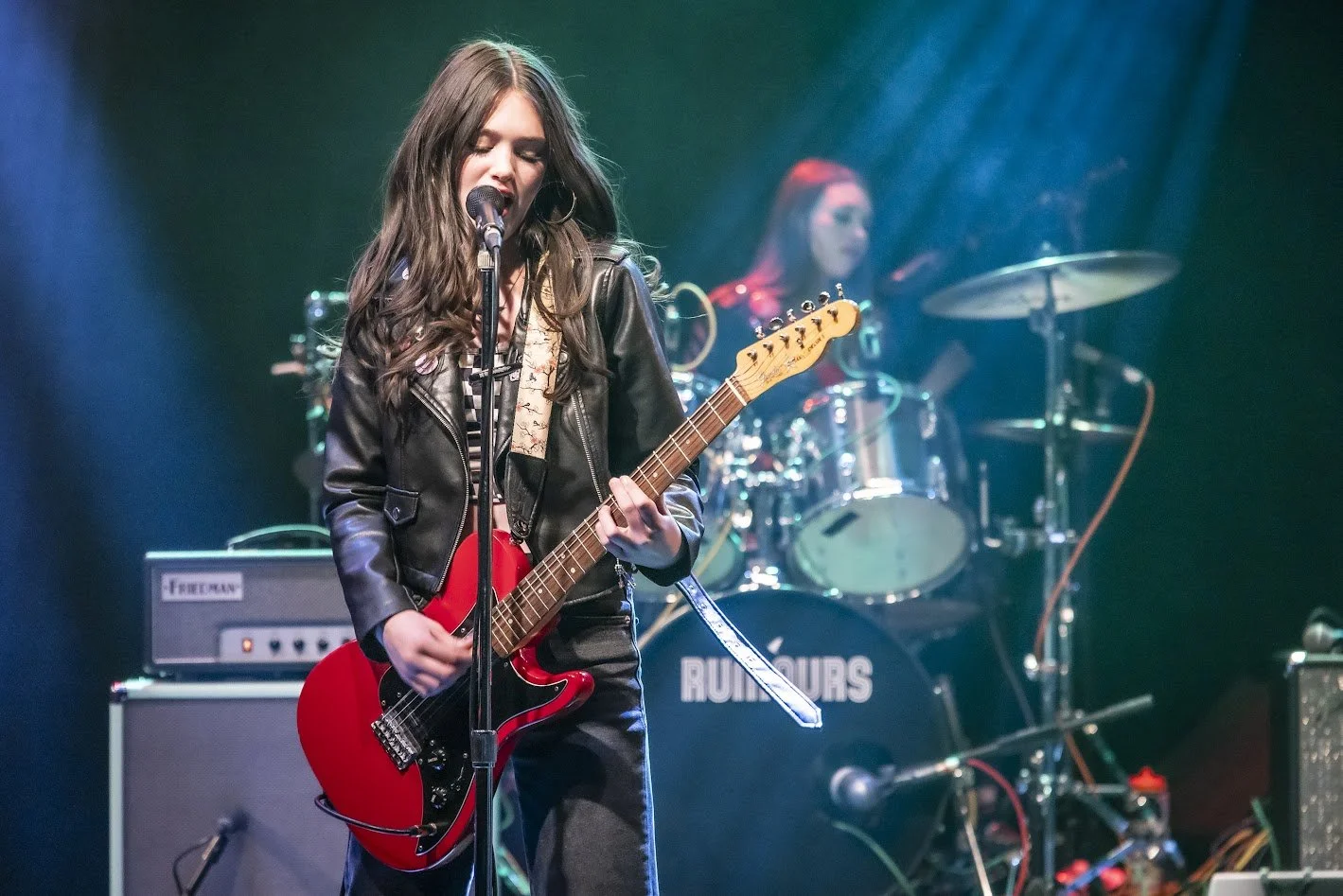Young woman playing electric guitar and singing into microphone on stage, with drummer in the background.