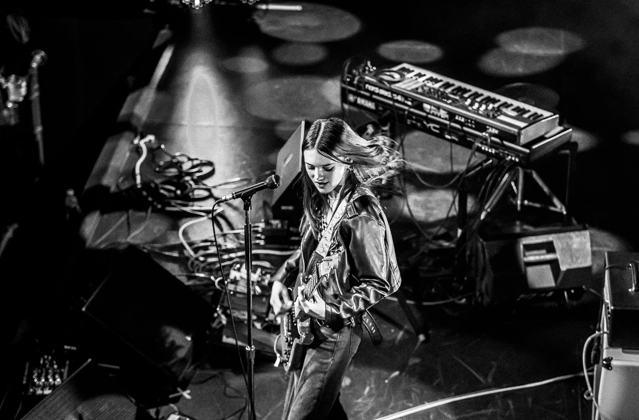 A young woman with long hair playing an electric guitar on stage, with musical equipment around her, in black and white.