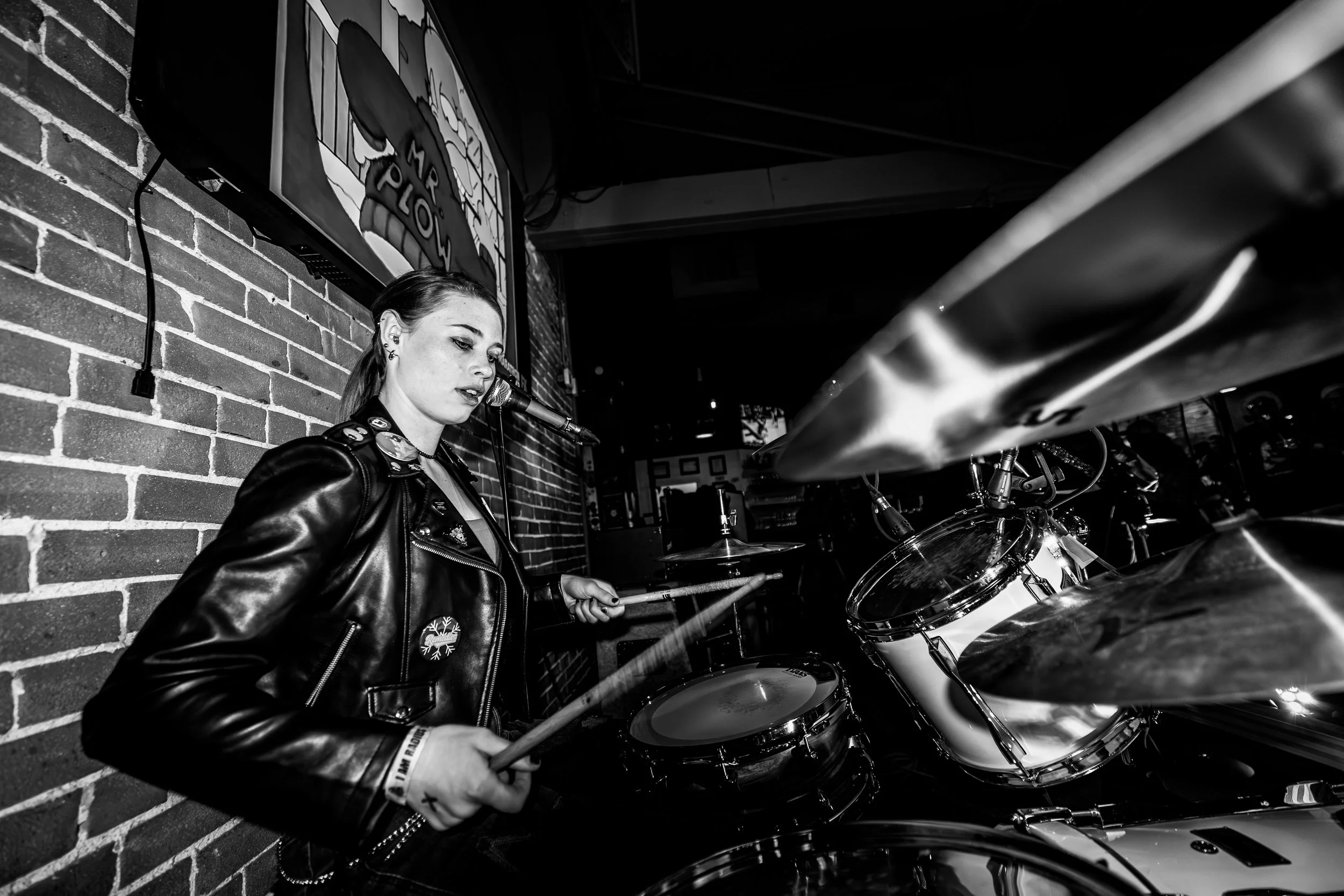 A young woman playing drums in a black-and-white indoor setting with a brick wall and a cartoon-themed sign in the background.