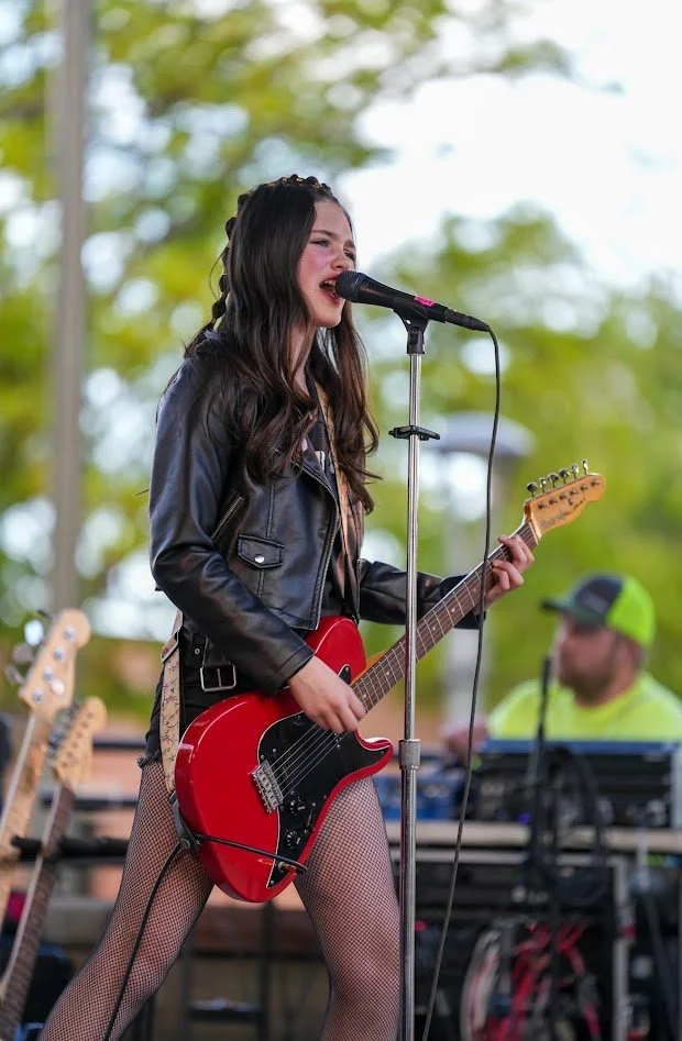 Young woman singing into a microphone while playing an electric guitar on stage outdoors, with a man in the background managing sound equipment.
