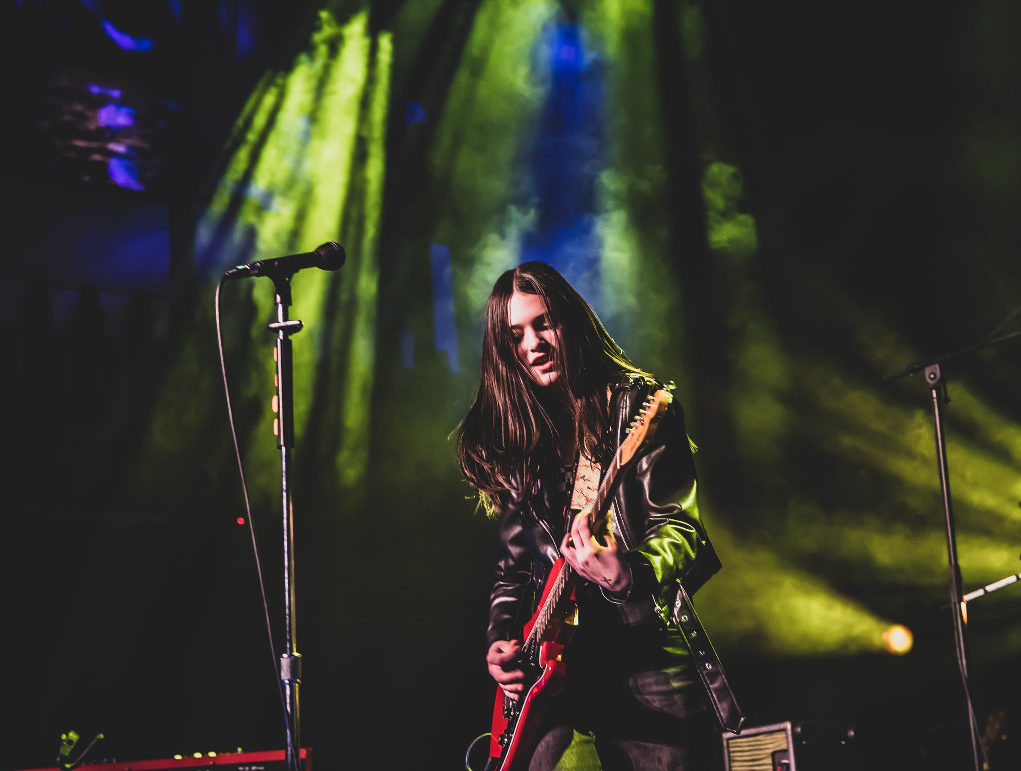 A young woman with long brown hair playing an electric guitar on stage in a dimly lit setting with green and blue stage lights projecting on the forest background.